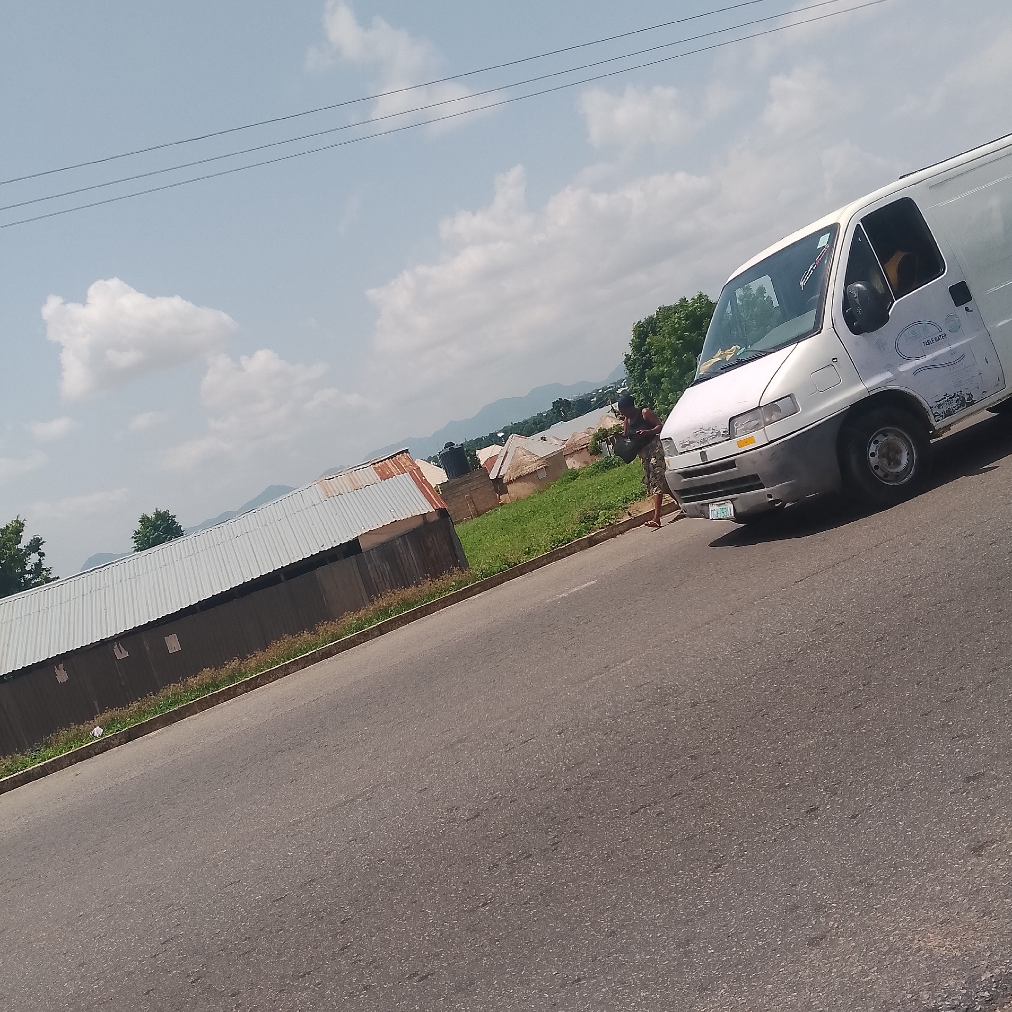 The image depicts a street scene in Jalingo, Nigeria, under a bright, partly cloudy sky, suggesting it is daytime. The foreground is dominated by a paved road that curves to the right. On the right side of the road, a white panel van is parked, with its driver's side door open and a person standing beside it. The person, wearing a dark sleeveless top and camouflage shorts, is holding a dark bag and appears to be walking away from the van. The van has some visible text on its side, including "TABLE WATER" in a stylized font. A license plate with the characters "DCA 793 AA" is visible on the front of the van.
To the left of the road, there's a row of buildings with corrugated metal roofs, some of which appear weathered. Beyond these structures, there are more buildings with thatched roofs, typical of a rural or semi-urban settlement. A water tank is visible on one of the buildings. The background features lush green grass and trees, with distant mountains forming the horizon. The overall atmosphere appears to be one of everyday life in a Nigerian town, with elements of both infrastructure and natural landscape. The lighting suggests it is likely mid-morning or afternoon.