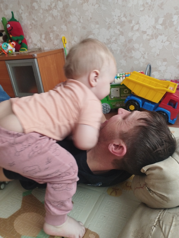 A baby stands on an adult lying on a mat. The baby wears a light peach shirt and patterned pink pants, and has bare feet. The adult wears a black shirt and rests their head on a pillow. Toys, including a truck, are scattered nearby on the mat and furniture. A cabinet and wall with a floral pattern are in the background.