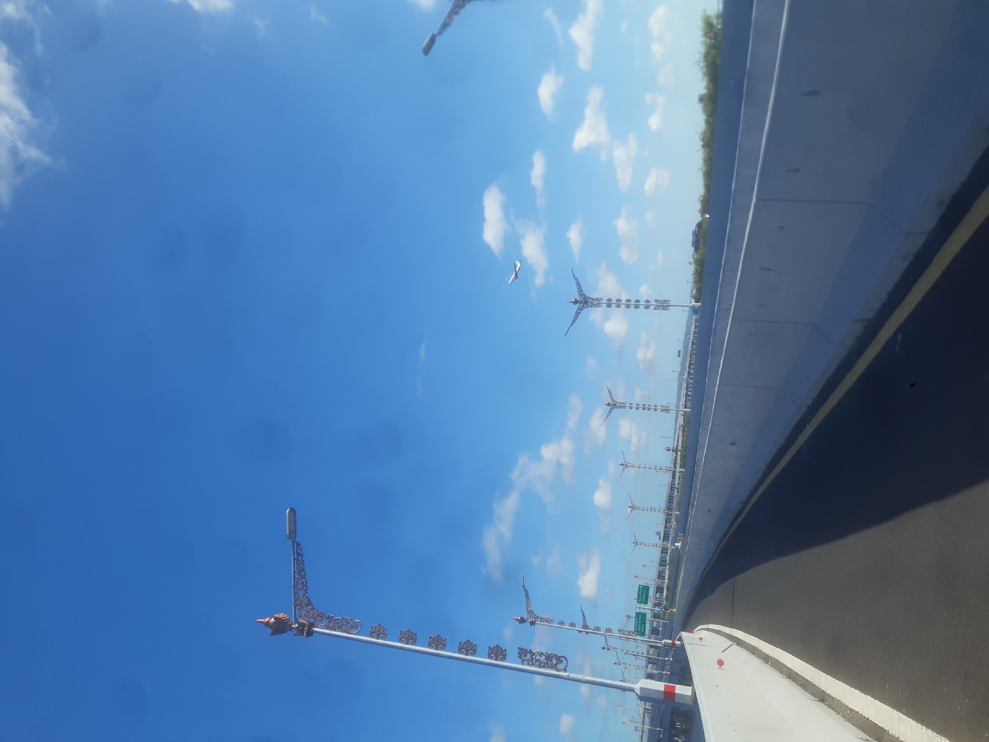 The image is taken from a low angle, looking up at a blue sky with scattered white clouds. A bridge or elevated road fills the lower portion of the frame, with a gray concrete barrier lining the side of the road. The road surface is dark asphalt. Ornate streetlights with decorative metalwork line the side of the road. In the distance, there are structures resembling windmills. A plane flies through the sky.
The location is likely the Bali Mandara Toll Road in Kuta, Indonesia, based on the distinct appearance of the streetlights and the windmills.