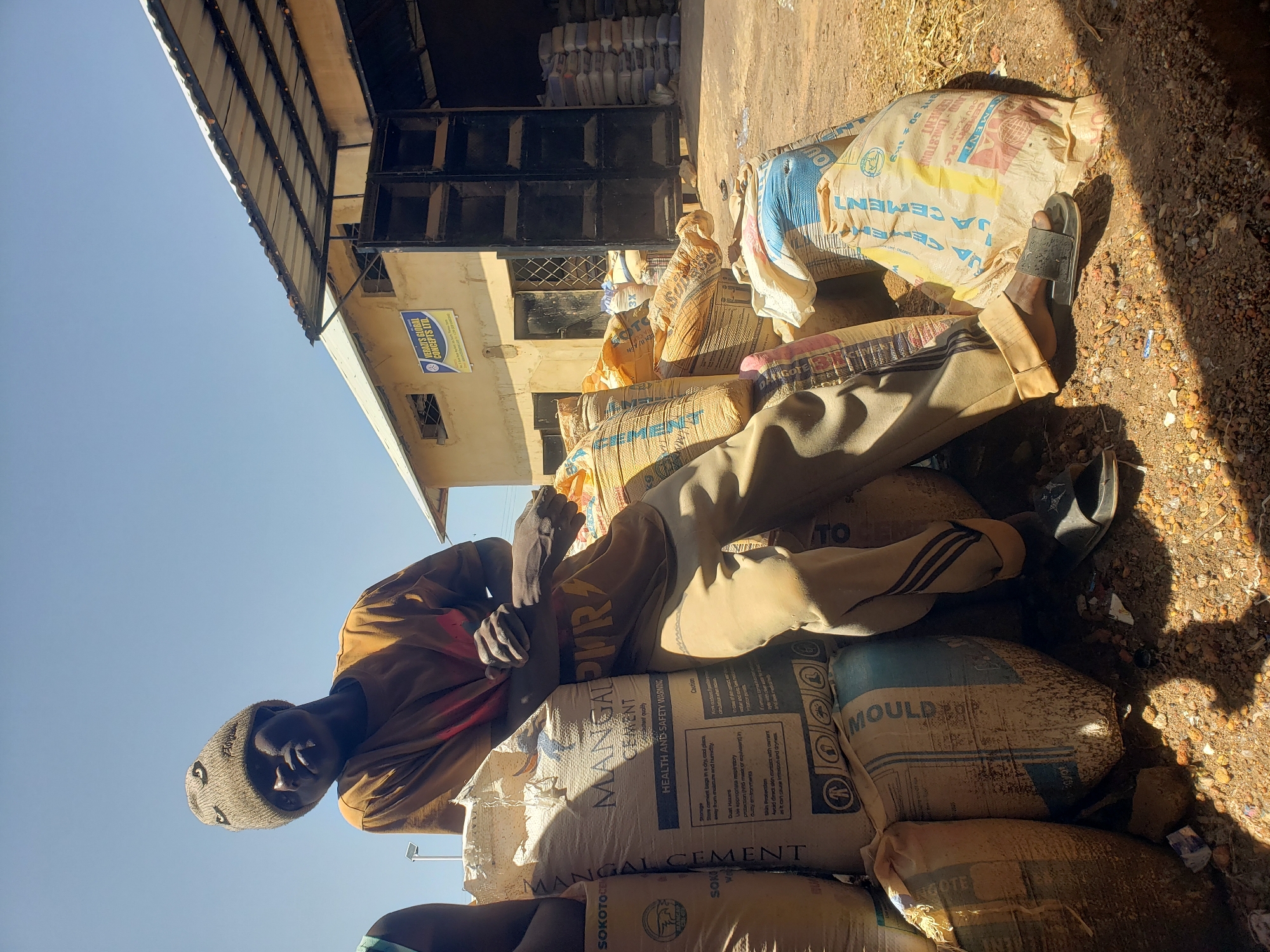 In Jos, Nigeria, under a clear blue sky, a young man sits casually amidst a large pile of cement bags. He is wearing a light brown beanie with an embroidered design and an orange t-shirt. His legs are crossed, with one foot resting on a black sandal. The cement bags are stacked high, some with visible labels including "SOKOTO CEMENT" and "MANGAL CEMENT". In the background, a building with a corrugated metal roof is visible, and more bags of cement are stacked outside its entrance. The ground is covered in reddish-brown soil, with scattered debris.