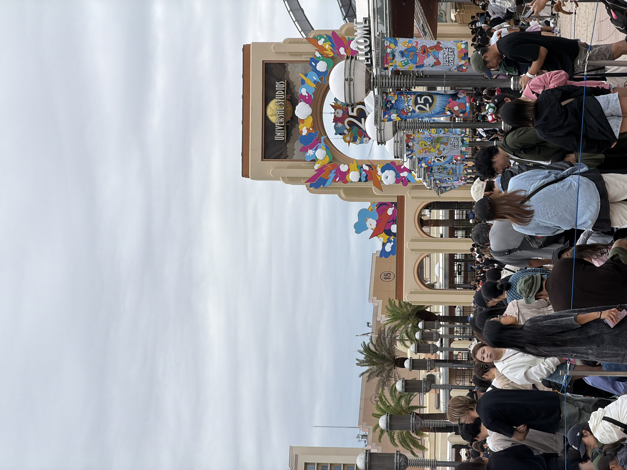 An outdoor, eye-level shot captures a bustling queue of people at the entrance of Universal Studios Japan in Osaka. The image is oriented vertically, showing the scene as if viewed from above and rotated 90 degrees counter-clockwise.
The main subject is a large crowd of individuals, predominantly adults, dressed in casual attire, forming lines to enter the theme park. Their backs are mostly towards the viewer, but some faces are visible, generally appearing patient.
In the background, a prominent architectural structure serves as the park's entrance. A large, ornate sign reads "UNIVERSAL STUDIOS JAPAN" on a building facade. Below this, a circular decorative element features colorful, cloud-like and flame-like shapes, surrounding a large number "25", indicating an anniversary celebration. To the right of the entrance, vertical banners featuring characters like those from "SESAME STREET" and "MINIONS" are visible, also displaying "UNIVERSAL STUDIOS" and the number "25." Another building segment has the number "(18)" inscribed.
The foreground is dominated by the queue lines, marked by metal stanchions and ropes. The people are densely packed, suggesting a busy day. In the middle ground, architectural elements typical of a theme park are seen, including buildings with arched windows and palm trees, adding to the theme park ambiance. The sky is overcast and bright, indicating daytime under cloudy conditions. Visible text includes "UNIVERSAL STUDIOS JAPAN", "WELCOME", "SESAME STREET", "MINIONS", and the number "25" repeated on various signs and banners.