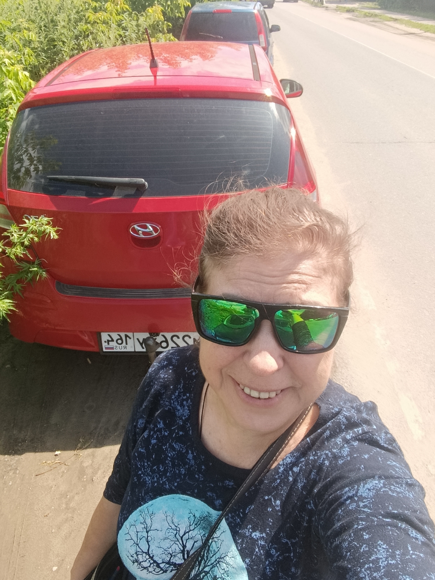 This media file is a selfie taken outdoors, likely in Orekhovo-Zuyevo, Russia. The main subject is a woman with brown hair, wearing sunglasses with green reflective lenses and a dark blue t-shirt with a stylized tree and moon graphic on the front. She is smiling, suggesting a pleasant mood.
Behind her, a red car is parked, partially obscuring the background. The car has a Hyundai logo on its rear. A license plate is visible on the car's bumper, showing Cyrillic characters. The car is parked on the side of a paved road, with a patch of dirt and green foliage next to it. In the background, more cars are visible on the road, and dense green bushes and trees line the opposite side.
The lighting indicates it is daytime, with bright sunlight casting shadows. The overall scene suggests a casual, possibly roadside stop during a trip or outing. The weather appears to be warm and sunny, conducive to outdoor activities.