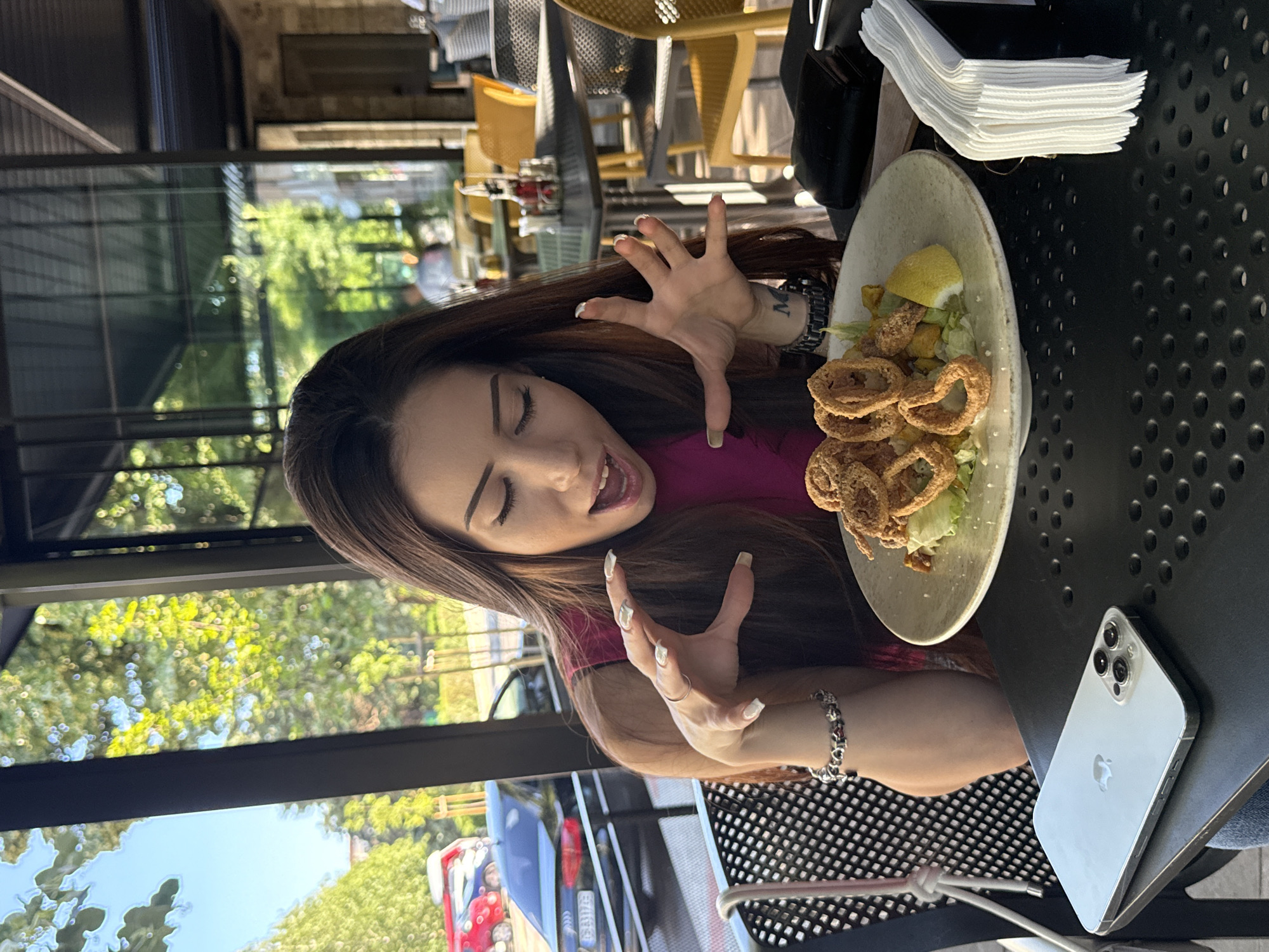 A young woman with long, dark hair is seated at an outdoor table, her mouth open in a surprised or excited expression. She is looking down at a plate of food in front of her, which appears to be calamari rings served over a salad with a slice of lemon. Her hands are raised with her fingers spread, as if reaching for or reacting to the food. She is wearing a pink top and a bracelet on her left wrist. A smartphone and a napkin holder are on the table. The background shows other tables and chairs, suggesting a restaurant or cafe setting. Trees and a building are visible through a glass-enclosed area behind her, with cars parked in the distance.