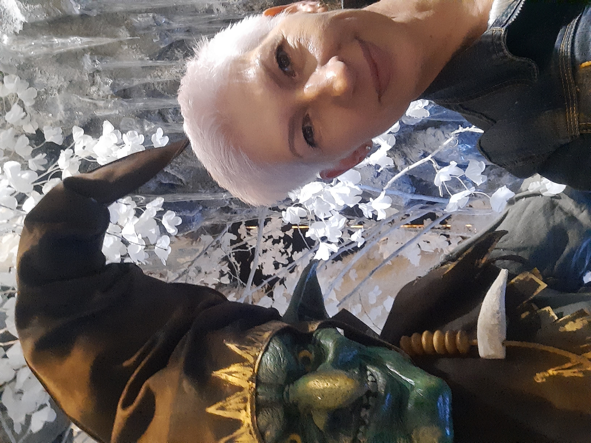 A woman with short, light-colored hair and fair skin is taking a selfie in front of a green, goblin-like figure wearing a pointed hat adorned with a gold crown. The background appears to be a decorative display with white flowers and icicle-like structures. The woman is wearing a dark denim jacket. The goblin figure has large yellow eyes, a prominent nose, and a wide grin, and is wearing a dark robe with gold accents and a string of wooden beads. The scene seems to be an indoor setting, possibly a themed attraction or event. It is impossible to determine the location within Minsk, Belarus from the image alone.
