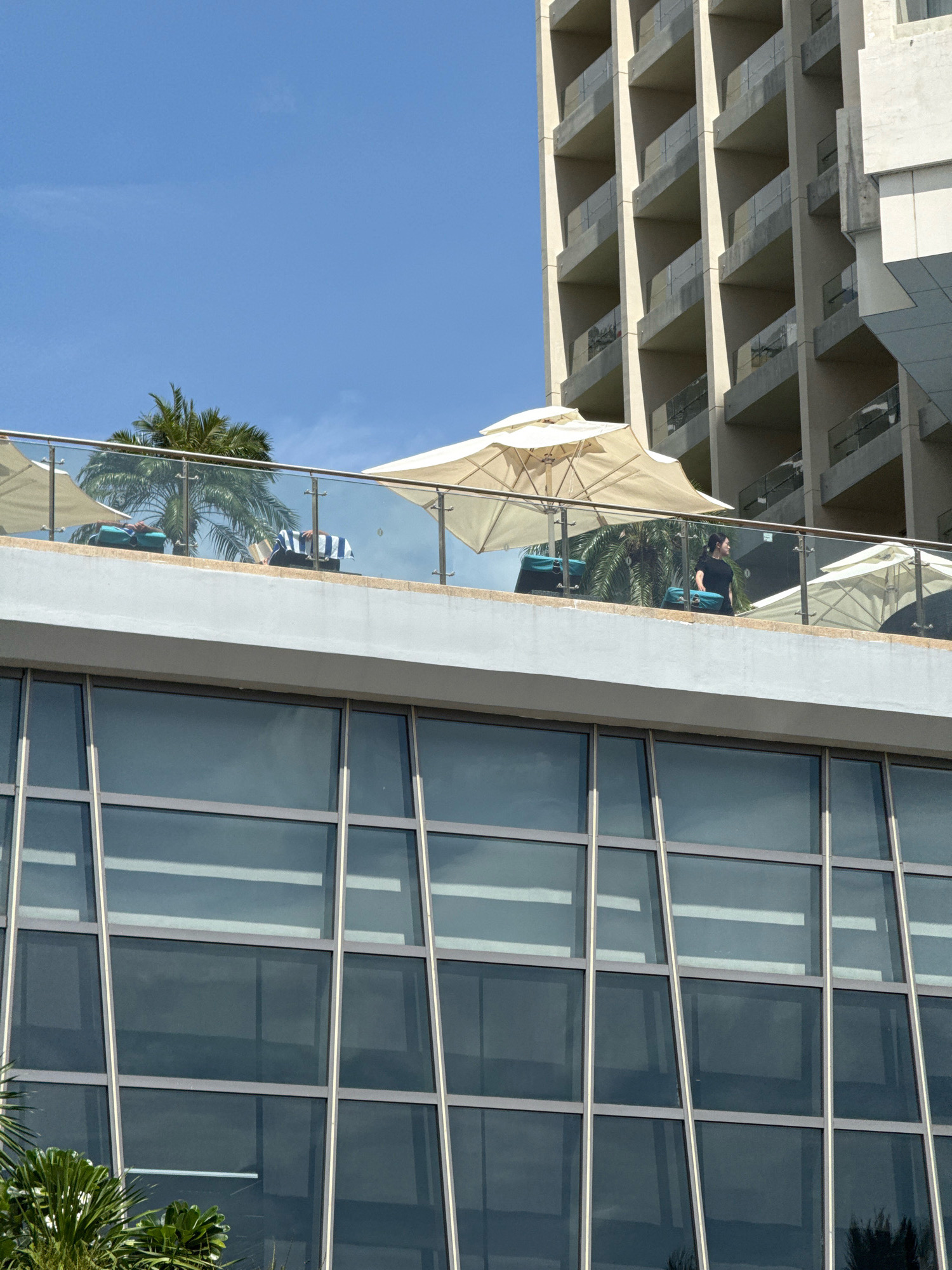 The image shows a rooftop terrace with lounge chairs and umbrellas, likely part of a hotel in Nha Trang, Vietnam. A person in a black shirt is standing by the glass railing, looking to the side. The terrace overlooks a modern building with multiple balconies. The sky is clear blue.