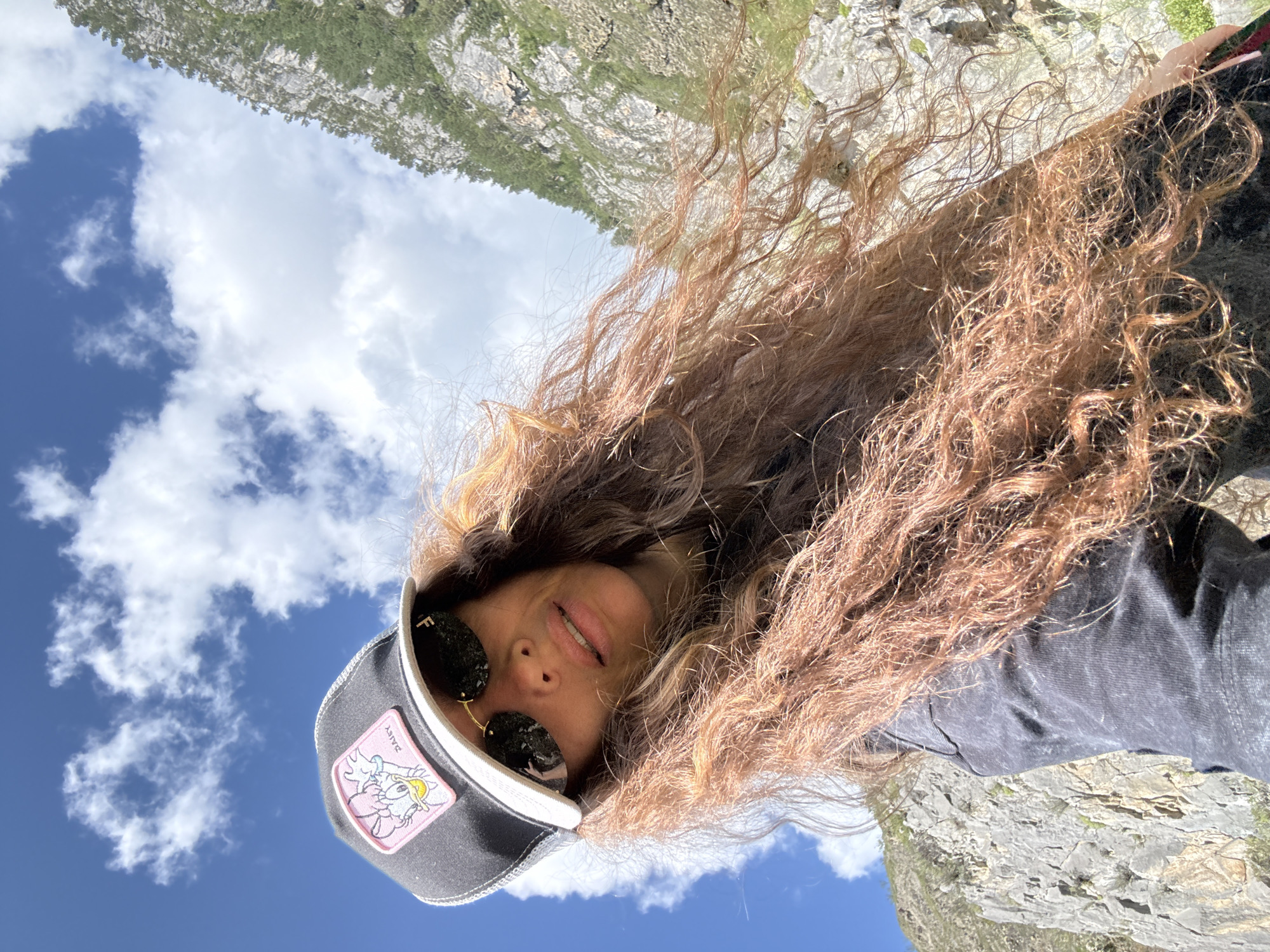 A person is taking a selfie outdoors, looking up at the sky. They are wearing a black baseball cap with a patch of Daisy Duck on it, sunglasses, and a dark grey t-shirt. Their long, curly, reddish-brown hair is blowing in the wind. The background shows a bright blue sky with white clouds, and a rocky mountainside with green trees. The image appears to have been taken in the vicinity of Chemal, Russia, a location known for its natural beauty and mountainous terrain.