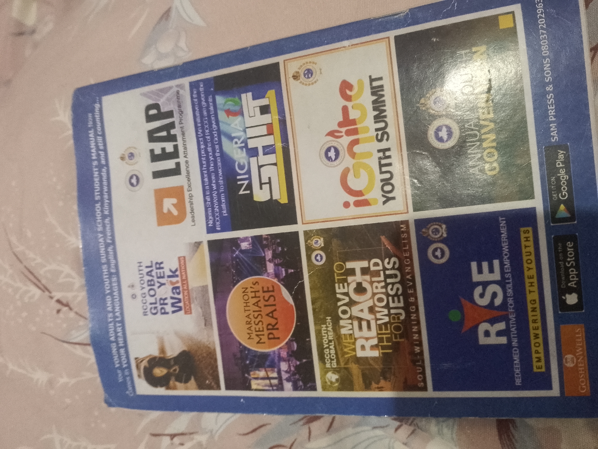 The image is a photograph of a brochure or flyer with multiple sections, each advertising a different event or program. The overall color scheme is blue and white with vibrant accent colors in the various sections. The brochure is angled and appears to be on a textured surface.
The top left section of the brochure has text that reads "THE YOUNG ADULTS AND YOUTHS SUNDAY SCHOO...". Below this, "COMES IN YOUR HEART LANGUAGES: English, French, Kinyarwanda, and still counting..." is visible. To the right of this text is a large white block with the word "LEAP" in bold black font with a red arrow pointing upwards. Below "LEAP" is "Leadership Excellence Attainment Programme". There is a description that states "Nigeria Shift is a talent hunt project (An initiative of the RCCGNYAYA) where the youths of RCCG are given the platform to showcase their God-given talents." Next to this text, the words "NIGERIA SHIFT" are prominently displayed in large, colorful letters.
To the right of "Nigeria Shift" is a section with the title "IGNITE YOUTH SUMMIT". The word "ignite" is stylized with flames and bright colors.
Further to the right, there is a section with the title "ANNUAL YOUTH CONVENTION". This section has a dark background.
Below the "LEAP" section is a section titled "RCCG YOUTH GLOBAL PRAYER WALK" with the subtitle "LOCATION ALL NATIONS". A portion of an image depicting people praying is visible.
Adjacent to the "GLOBAL PRAYER WALK" section is a section for "MARATHON MESSIAH'S PRAISE". This section features a circular orange logo with the text "MARATHON MESSIAH'S PRAISE".
To the right of the "MARATHON MESSIAH'S PRAISE" section is a section titled "WE MOVE TO REACH THE WORLD FOR JESUS". Below this title is "SOUL-WINNING & EVANGELISM".
The final section on the right features the word "RISE" in large, bold letters with an orange triangle icon. Below "RISE" it reads "REDEEMED INITIATIVE FOR SKILLS EMPOWERMENT" and "EMPOWERING THE YOUTHS". Below this, there is a QR code and options to download on the App Store, as well as a logo for "GOSHENWELLS".
The top right corner of the brochure has text that reads "SAM PRESS & SONS 08037202963". There is also a "GET IT ON Google Play" logo.