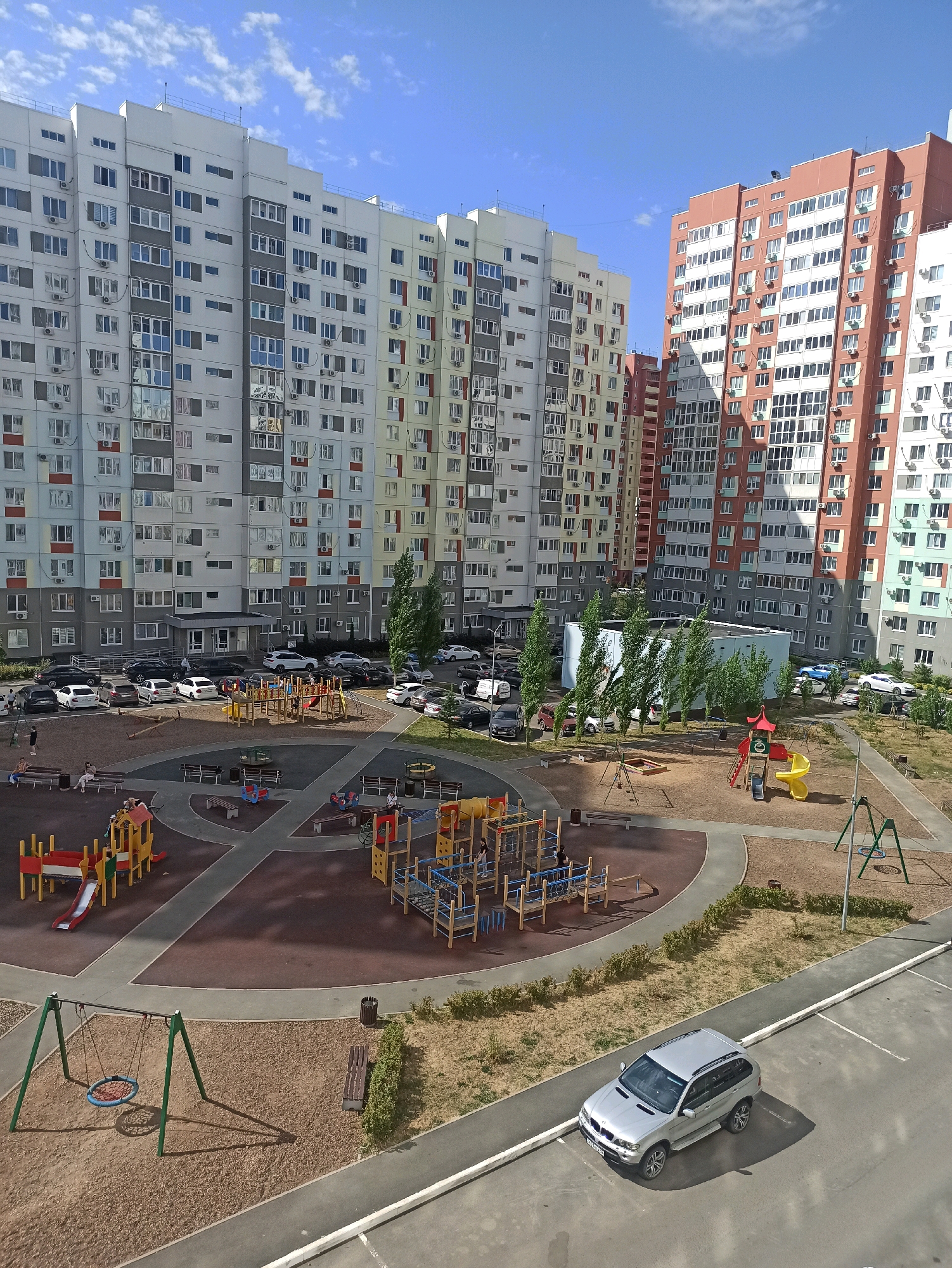The image depicts a residential courtyard with multiple high-rise apartment buildings and a playground. The buildings are modern, with varied architectural styles and colors, including white, yellow, and orange facades. The playground features slides, swings, climbing structures, and sandboxes, with some children and adults present. Several cars are parked in a designated lot adjacent to the courtyard. The scene is set against a clear blue sky with scattered clouds. The location is the residential complex "Aksakovsky", Orenburg, Russia.