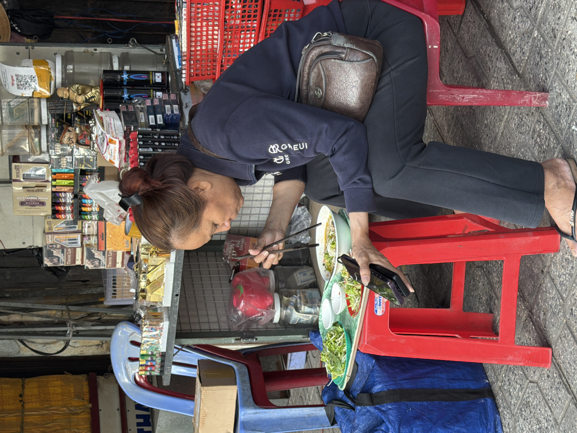 A Vietnamese woman sits on a red plastic stool and eats a bowl of noodles. She is dressed in a dark blue tracksuit and has a brown leather purse slung over her shoulder. In front of her is a tray with two small bowls of bean sprouts and a bowl of noodles, which she is eating with chopsticks. She is holding a black mobile phone in her left hand. The scene appears to be outdoors, possibly on a street in Nha Trang, Vietnam, with various goods displayed on shelves behind her, including lighters, packaged cigarettes, and some decorative figurines. The pavement is tiled. A blue tarp-covered object is visible to her left.
