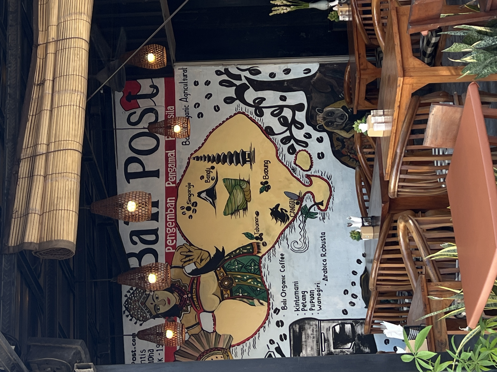 The image shows a sign for Bali Post, a cafe located in North Kuta, Indonesia. The sign features a map of Bali, with specific regions labeled, including Singaraja, Bangli, Tabanan, and Canggu. The sign also lists Bali Organic Coffee and Arabica Robusta as offerings. There is a mural featuring a traditional Balinese figure. Wicker lamps hang above the sign. Wooden tables and chairs are visible. Plants are also present, adding to the ambiance.