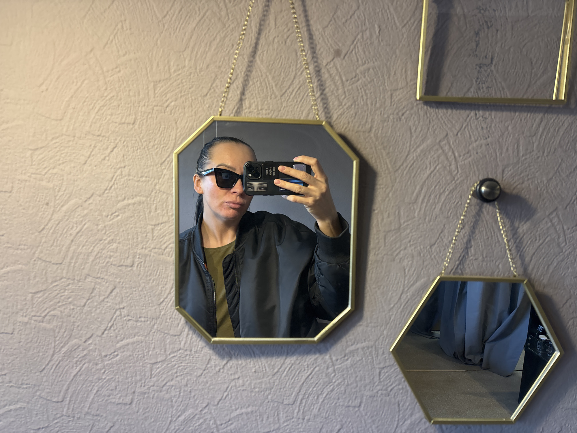 The image is a selfie taken in a mirror. The person in the reflection is wearing sunglasses, a black bomber jacket, and holding a phone. Behind the main mirror are two other mirrors hanging on a textured grey wall. The location cannot be determined based solely on the provided image. The city of Chaikovsky, Russia, cannot be ascertained from the visual information provided.