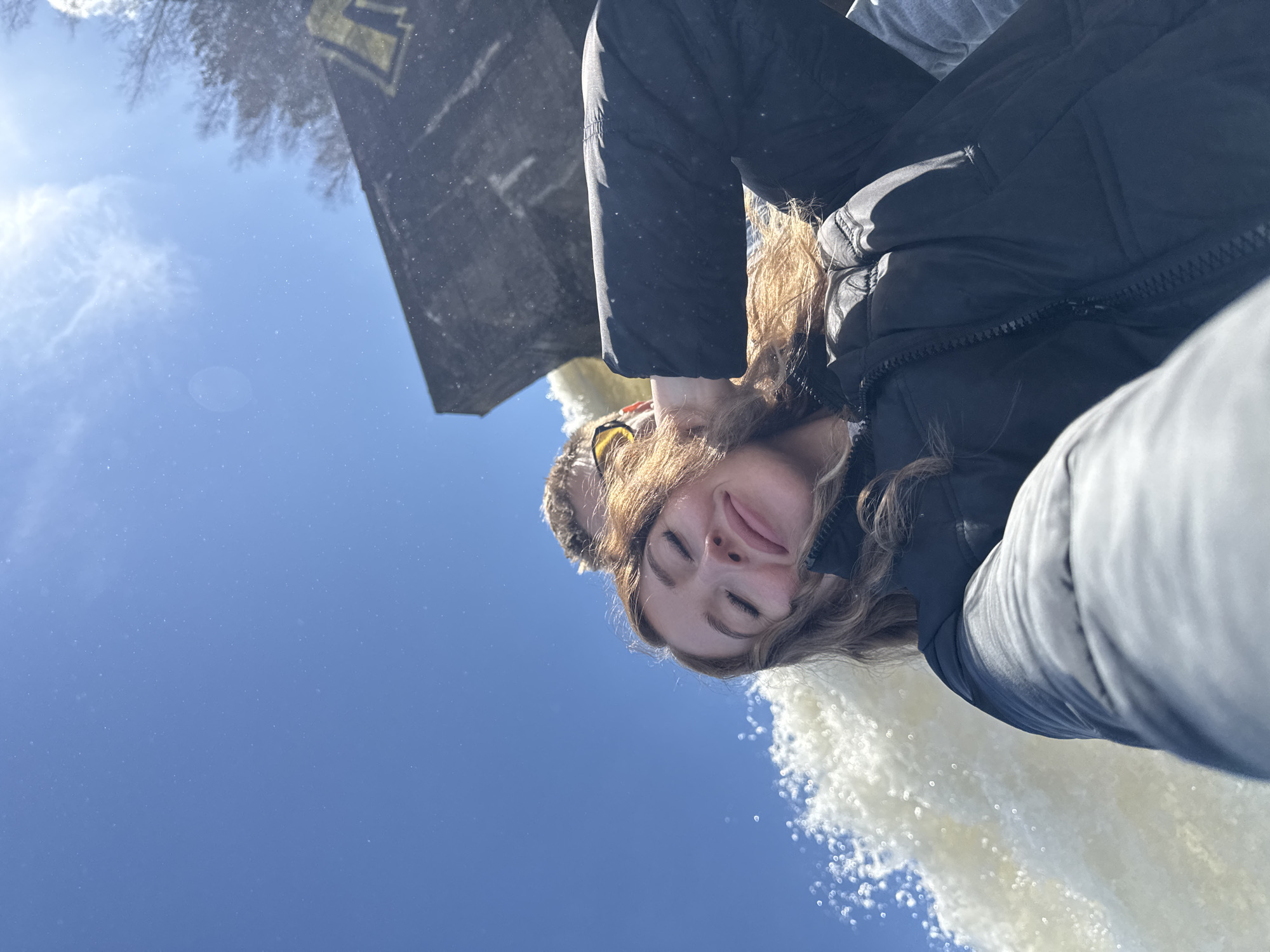 This outdoor photograph features a young woman in a close-up, selfie-style shot, set against a bright blue sky. She is seen from a low angle, looking upwards with her eyes closed and a gentle smile, suggesting a moment of peace or enjoyment. Her long, wavy light brown hair frames her face, and she is dressed in a dark, possibly black, puffer jacket and what appears to be a furry hat or earmuffs. A portion of a grey garment, likely belonging to another person, is visible behind her shoulder.
To her right, a large volume of white, foamy water spray, indicative of a waterfall or powerful fountain, adds dynamic texture to the scene. The background is dominated by a clear, azure sky with faint wisps of white clouds, accentuated by a sun flare. Numerous tiny white specks are scattered across the blue expanse, possibly water droplets from the spray or dust on the camera lens.
In the upper left corner, a dark, angular architectural structure or building is partially visible. It bears a prominent yellow or gold emblem, which resembles a stylized letter like an "M" or "W." Bare tree branches are also discernible against the sky near this structure. The strong sunlight indicates it is daytime. The scene captures a serene moment at a scenic location, potentially in Noviy, Russia, given the provided context.