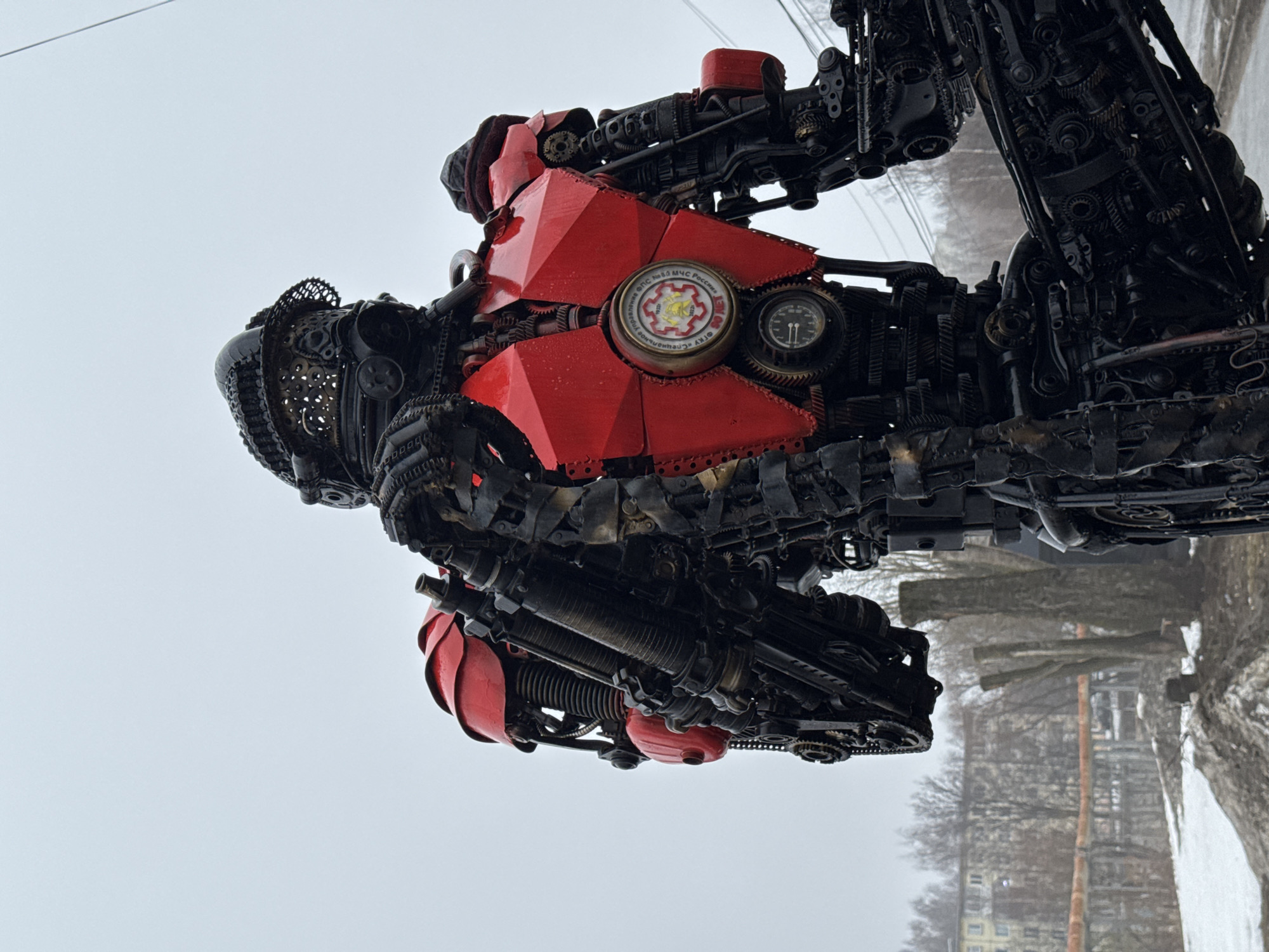 The image shows a large, complex metal sculpture in the city of Votkinsk, Russia. The sculpture appears to be a fantastical machine or robot, constructed from various metal components, including gears, chains, and pipes. Some parts of the sculpture are painted red, while others are dark and metallic. A circular badge featuring an emblem with text in Russian is visible on the sculpture's front. The sculpture is located outdoors, with a view of trees, buildings, and a cloudy sky in the background. The ground appears to be covered with snow or ice.