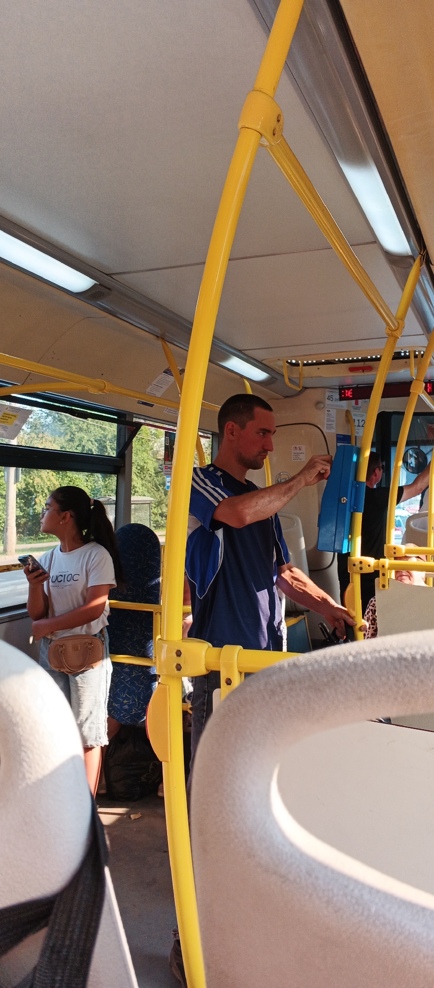 The image depicts the interior of a public bus in Izhevsk, Russia. In the foreground, a man wearing a blue and white striped t-shirt is seen interacting with a ticket validation machine. He is standing and appears to be holding a ticket or card. To his left, a young woman with dark hair pulled back in a ponytail is standing and holding a smartphone in her left hand. She is wearing a white t-shirt with text, denim shorts, and a small tan crossbody bag. She is looking towards the left side of the bus. The bus interior is characterized by yellow handrails and poles, beige ceiling panels with fluorescent lighting, and a dark floor. Outside the bus windows, greenery and trees are visible, suggesting the bus is in transit on a sunny day. The overall scene is a typical snapshot of daily life on public transportation.