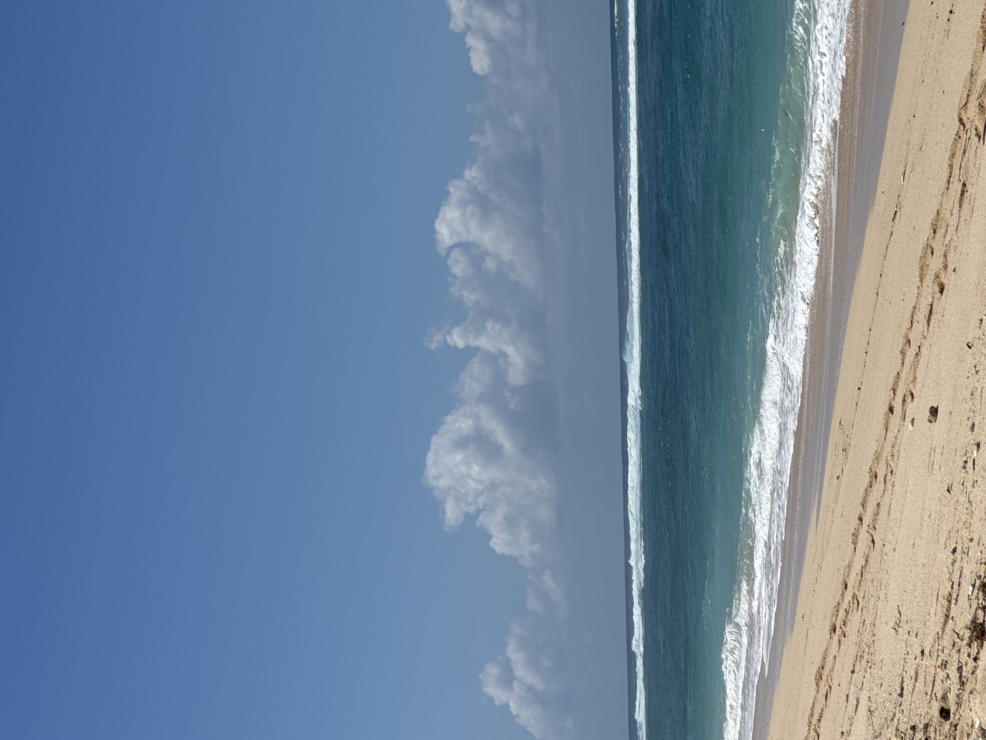 The image depicts a beach scene in Kuta Selatan, Indonesia. The foreground is dominated by a sandy beach with footprints and scattered pebbles. The ocean stretches across the middle ground, exhibiting shades of deep blue and turquoise, with white waves crashing near the shore and a line of larger waves breaking further out. The horizon is marked by the meeting of the ocean and the sky. The sky above is a clear, vibrant blue, punctuated by several fluffy white clouds. The scene is natural and serene, with no people or man-made structures visible.