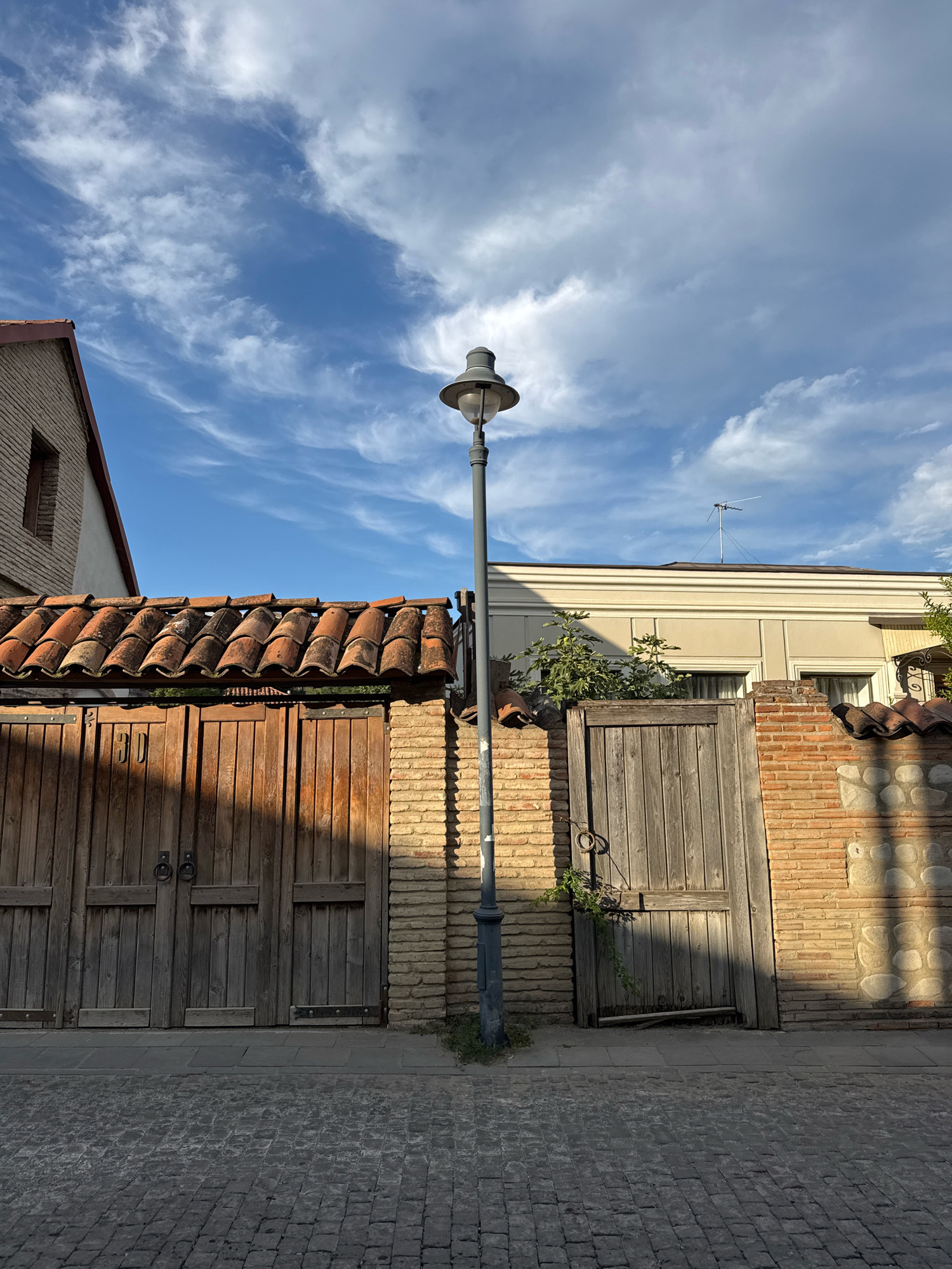 The image depicts a street scene in Telavi, Georgia, featuring traditional architecture. In the foreground, a cobblestone street leads to a weathered wooden gate, flanked by brick walls. Above the gate, a tiled roof juts out. To the left, another building with a similar tiled roof and a single window is visible, constructed from light-colored bricks. A tall, grey lamppost stands prominently in the center of the frame, casting a shadow onto the street. The background reveals a building with cream-colored walls and windows, topped by a television antenna. The sky above is a vibrant blue, streaked with wispy white clouds.