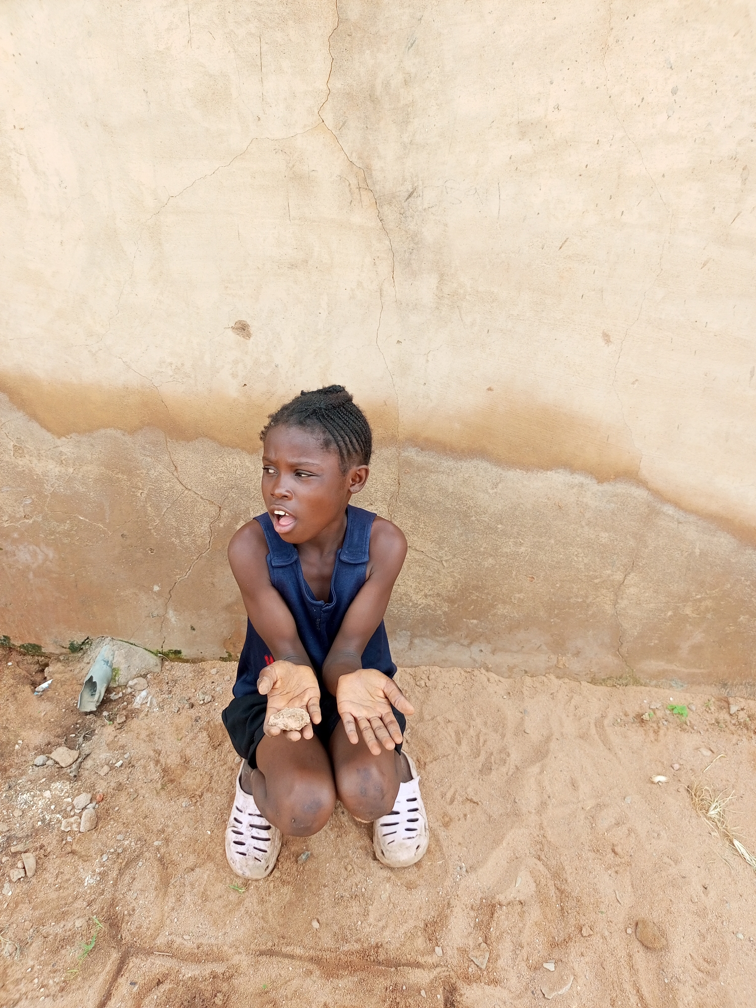 In the city of Tempure, Nigeria, a young girl of African descent kneels on a sandy ground in front of a beige, cracked plaster wall. She is wearing a dark blue sleeveless dress and light-colored shoes. Her mouth is open, and she appears to be looking to her left with a surprised expression. Her hands are open and held out in front of her, with one hand containing a small piece of dirt or stone. The environment is dry and dusty, with scattered debris on the ground, including a broken pipe. The lighting suggests it is daytime, with the sun casting shadows.