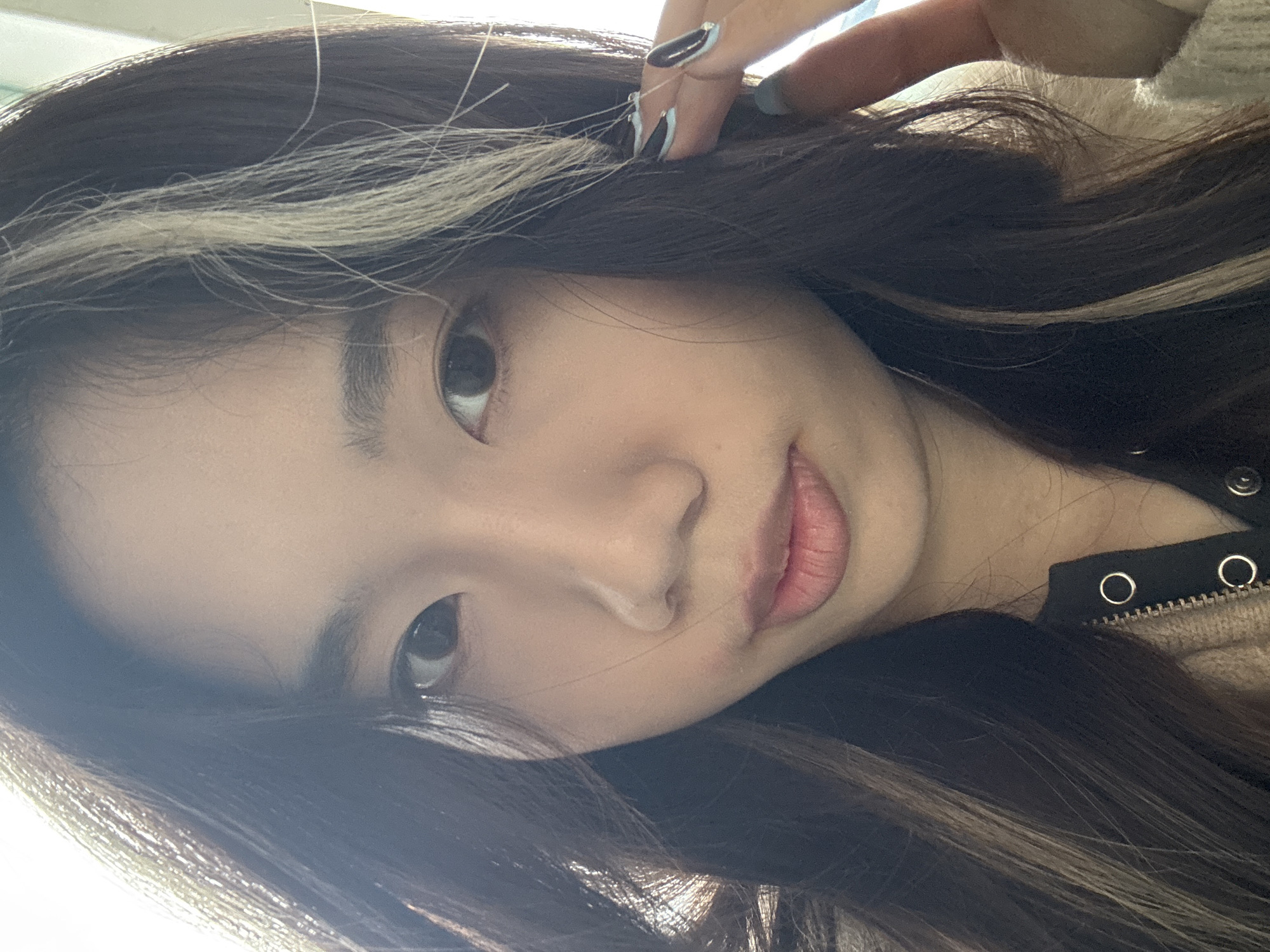 A close-up portrait shows an individual, likely female, of East Asian appearance, facing the camera directly. The subject has dark eyes, dark brown hair with prominent light blonde or light brown highlights framing the face, and light pink lips slightly parted. She is wearing a light beige or brown knitted outer garment with a zipper and silver snap buttons over a dark undershirt. Her left hand is raised to the side of her head, with fingers touching or near her hair. The fingernails on this hand feature a black and light blue French tip design, and one nail has a small silver embellishment. The background is brightly lit and out of focus, providing no specific environmental details. The exact location within Nan'an District, China, cannot be determined from the visual information provided.