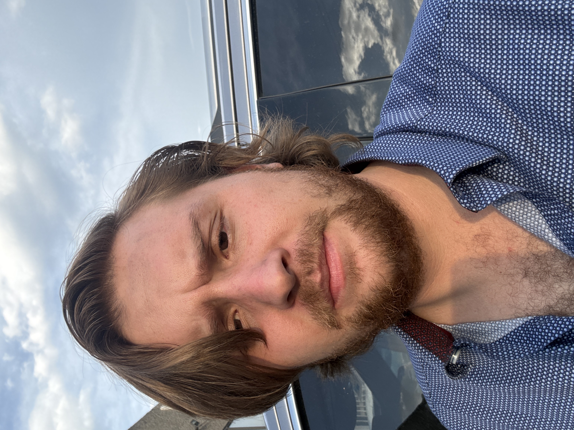 A close-up, slightly angled shot features a man with brown hair and a beard, wearing a blue patterned shirt. He is standing outdoors. The background includes a vehicle with a reflective surface showing a cloudy sky. The image appears to have been taken during daylight. Without additional information, it is impossible to determine the location in Cuautitlán Izcalli, Mexico.