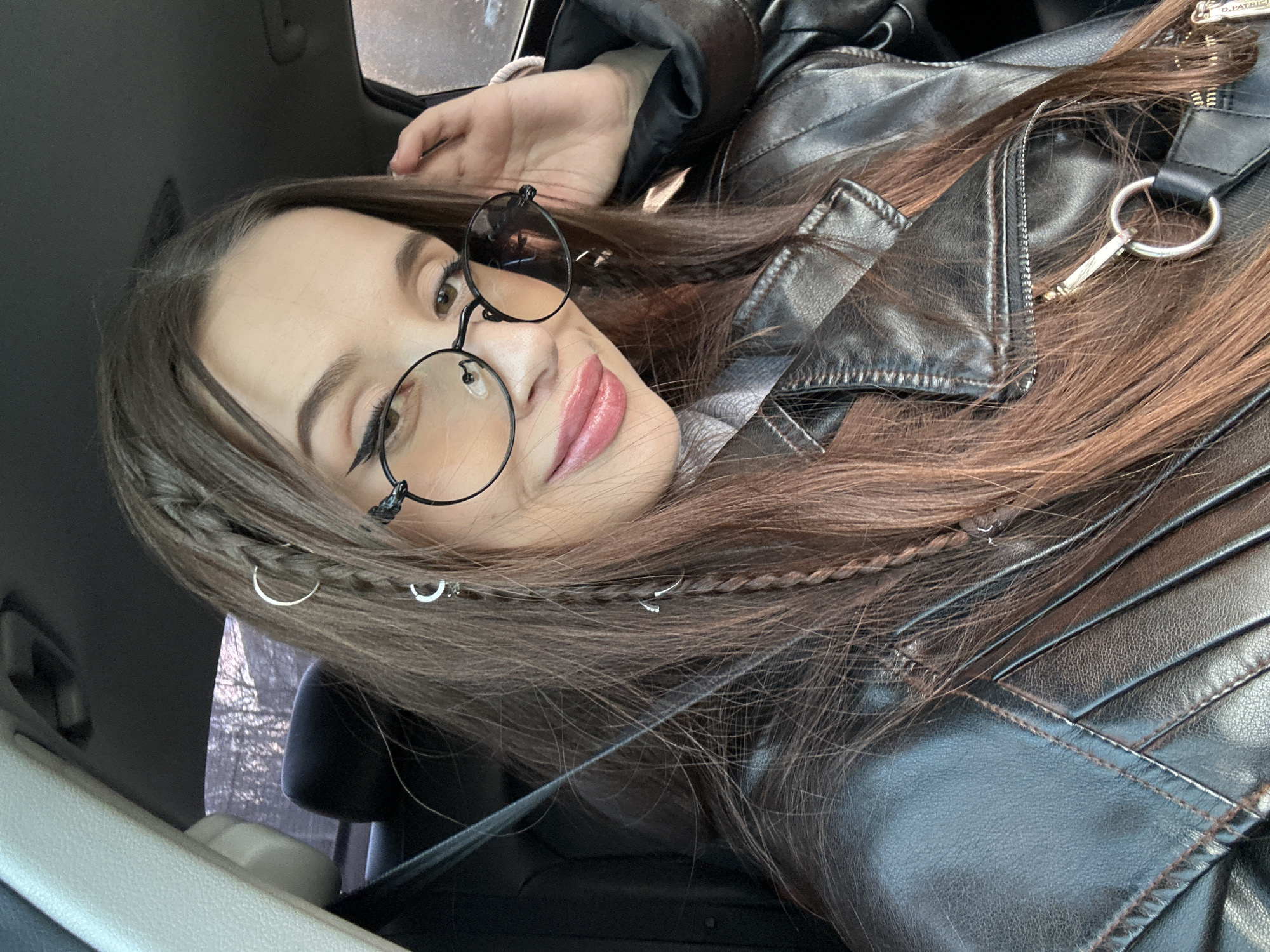 A portrait-oriented photograph displays a young woman looking directly at the camera with a subtle smile. She is positioned as if reclining, tilted approximately 90 degrees clockwise from an upright stance. She has long, straight brown hair, which features small braids adorned with several silver-colored rings and clips.
The woman wears round, black-framed glasses and has visible winged eyeliner. Her lips are painted a soft pink or mauve shade. She is dressed in a black faux leather jacket, which exhibits zippered details and a padded or quilted design on the shoulders. A silver D-ring and a key-like charm are attached to a zipper pull on her jacket, where the text "O.PATRIC" is faintly discernable.
The setting is an interior space, likely a car, indicated by light-colored plastic trim and dark upholstery visible in the background. A blurred reflection in a window suggests an outdoor environment, possibly with trees or distant architecture. The lighting indicates it is daytime. The location context for this image is Yekaterinburg, Russia.