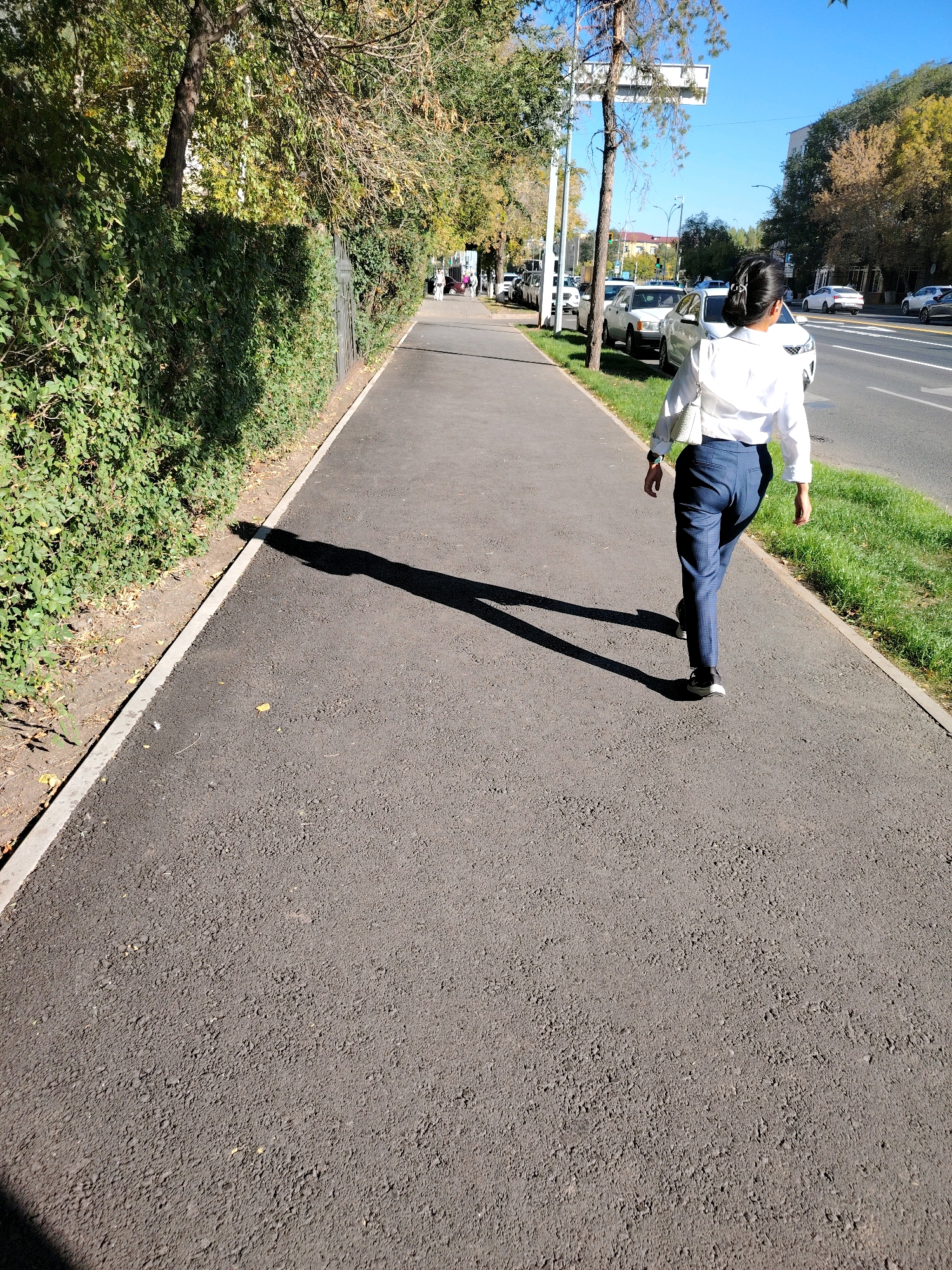 A woman wearing a white blouse and plaid blue pants walks away from the camera on a paved pedestrian path in Astana, Kazakhstan. She carries a white purse on her left shoulder. To her left is a dense green hedge and trees with autumn foliage. To her right, beyond a grassy area, is a multi-lane road with several cars and apartment buildings in the background under a clear blue sky. The sun casts a long shadow of the woman across the path.