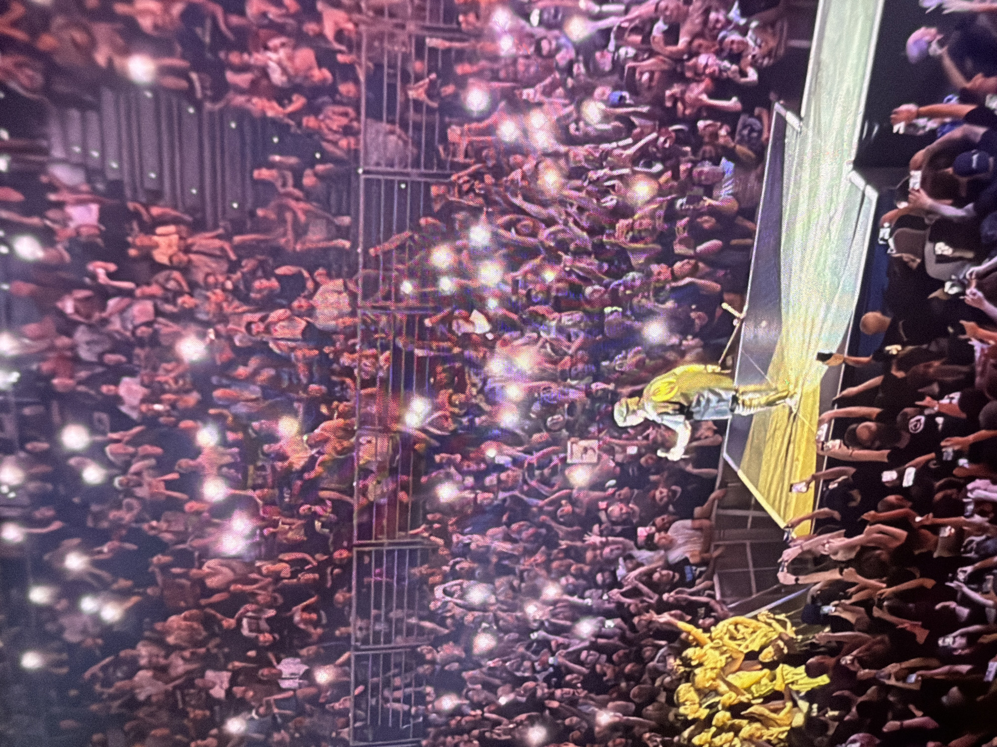 A concert is taking place in a stadium with a large audience in attendance. The audience members are visible in tiered seating, and many are holding up their mobile phones with their flashlights on, creating a sea of lights. In the foreground, on a brightly lit stage, a performer is standing and facing the audience. The performer is wearing a dark outfit with some lighter colored accents. The overall atmosphere appears energetic and celebratory.