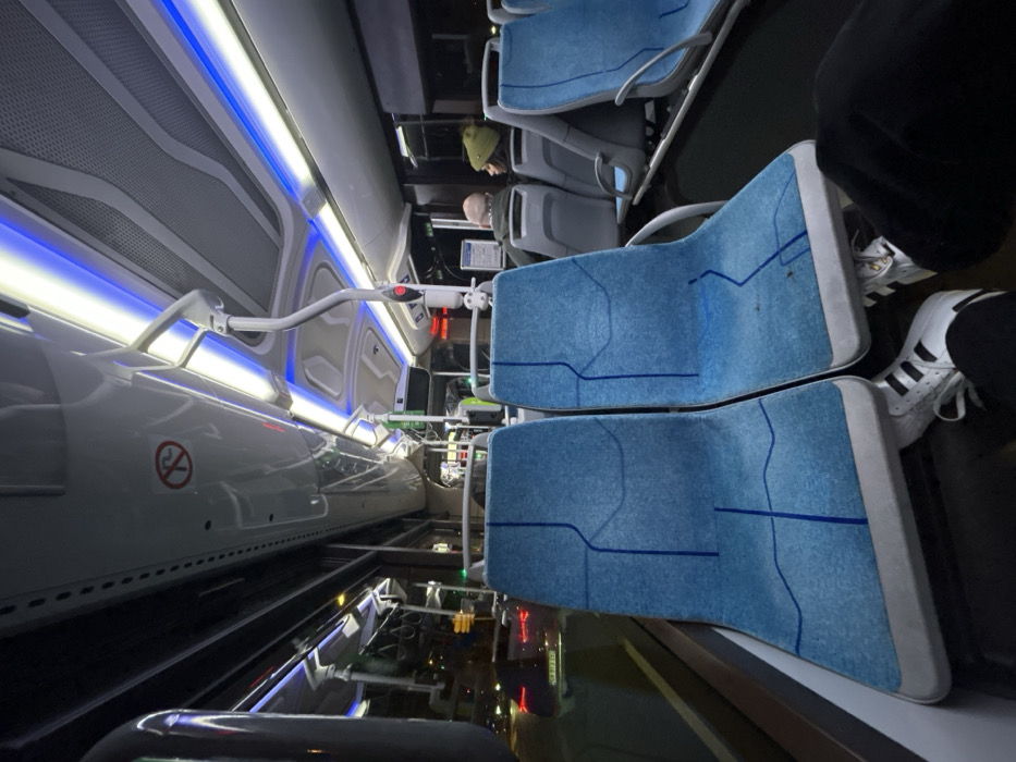 The image captures the interior of a modern public transport vehicle, likely a bus or tram, taken from a sideways perspective (rotated 90 degrees clockwise). The scene shows a brightly lit, clean, and futuristic-looking cabin, suggesting it is evening or night due to the reliance on artificial lighting and reflections from outside.
On the right side of the image, several rows of light blue upholstered seats are visible. These seats feature a subtle, darker blue geometric or abstract line pattern. The seat backs and bases are framed with light grey or white material. In the immediate foreground, a person's lower legs and feet are visible, wearing dark trousers and white Adidas sneakers with black stripes, resting near an empty seat. Further down the aisle, two other individuals are seated. One person, seen from behind, wears a green or olive-colored beanie/hat. Beside them, another person's head, possibly balding, is partially visible.
The left side of the image showcases the vehicle's interior wall and ceiling. Sleek, light grey or white panels line the area, illuminated by prominent horizontal LED strip lighting. These lights emit both cool white and blue hues, casting a distinct glow throughout the cabin. Above the seating area, a white, curved grab bar extends from the wall towards the center, ending in a handle with a red button or indicator. Higher up, another similar but shorter grab bar is present. The upper wall panels feature a perforated or mesh design.
Near the bottom left of the image, on a lower side panel, a "no smoking" symbol is clearly visible: a red circle with a diagonal line over a cigarette icon. The floor of the vehicle appears dark and highly reflective, mirroring the internal lights and showing blurred reflections of what might be external city lights or other vehicles passing by, reinforcing the impression of travel at night. The overall impression is one of modern design, efficiency, and cleanliness within an urban transport setting.