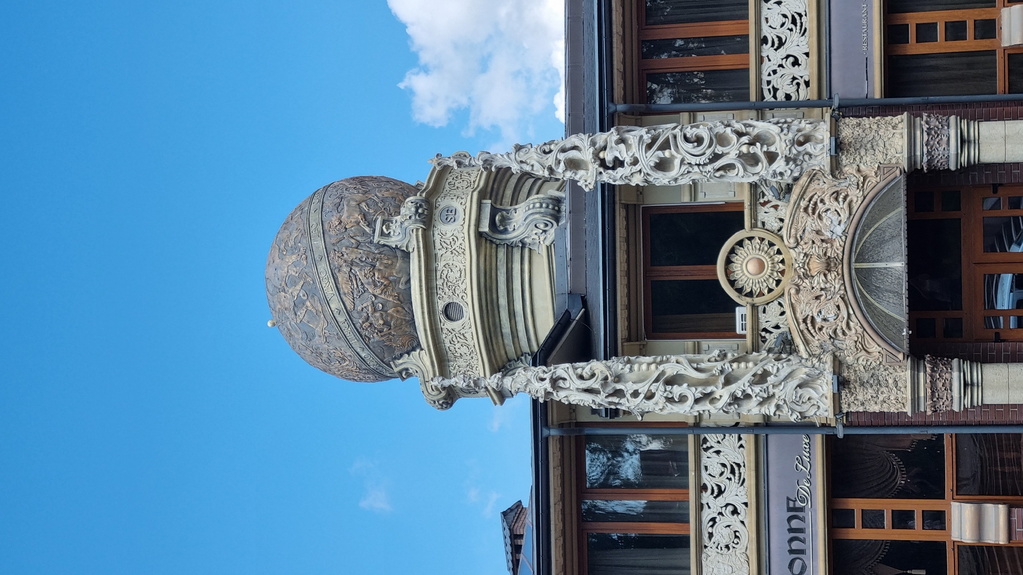 The image shows the ornate facade of the "De Luxe" restaurant in Chernivtsi, Ukraine. The building features a prominent domed structure on its roof, decorated with intricate carvings depicting zodiac signs and mythological figures. The facade is adorned with elaborate stucco work, including decorative columns, floral motifs, and a sun-like emblem above the entrance. The windows are framed with dark wood, and the building's entrance is marked by an arched doorway with a glass canopy. A sign with the text "SONNE De Luxe" is visible on the left side of the building. The sky above is a clear blue with a few scattered white clouds. The image is a close-up shot, focusing on the architectural details of the restaurant.