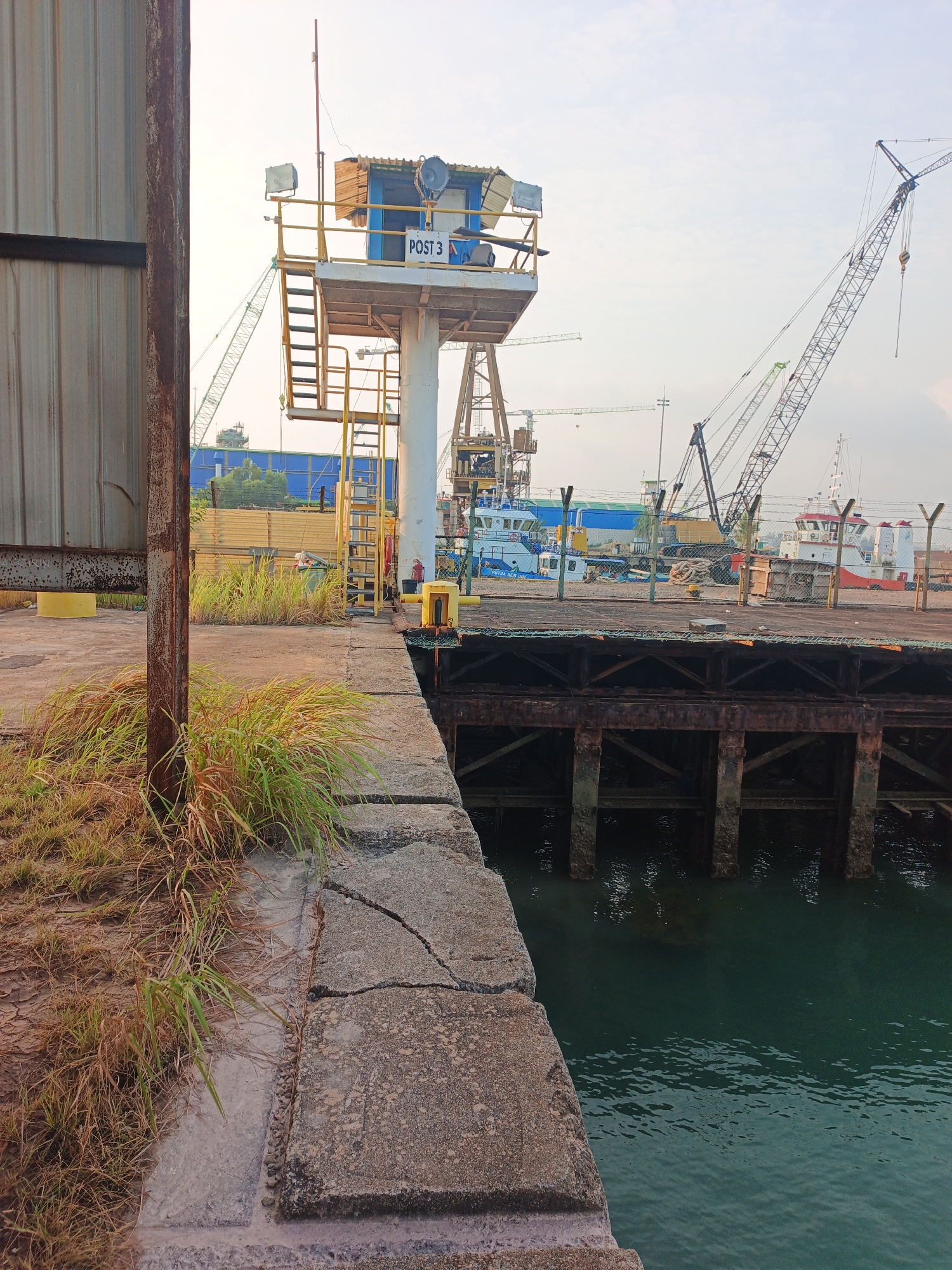 This image depicts a dockside area with a prominent "POST 3" sign on a raised platform. Several cranes are visible in the background, suggesting a port or industrial area. The foreground shows a concrete walkway with overgrown grass and a view of the water. The scene appears to be located in the city of Batam, Indonesia.