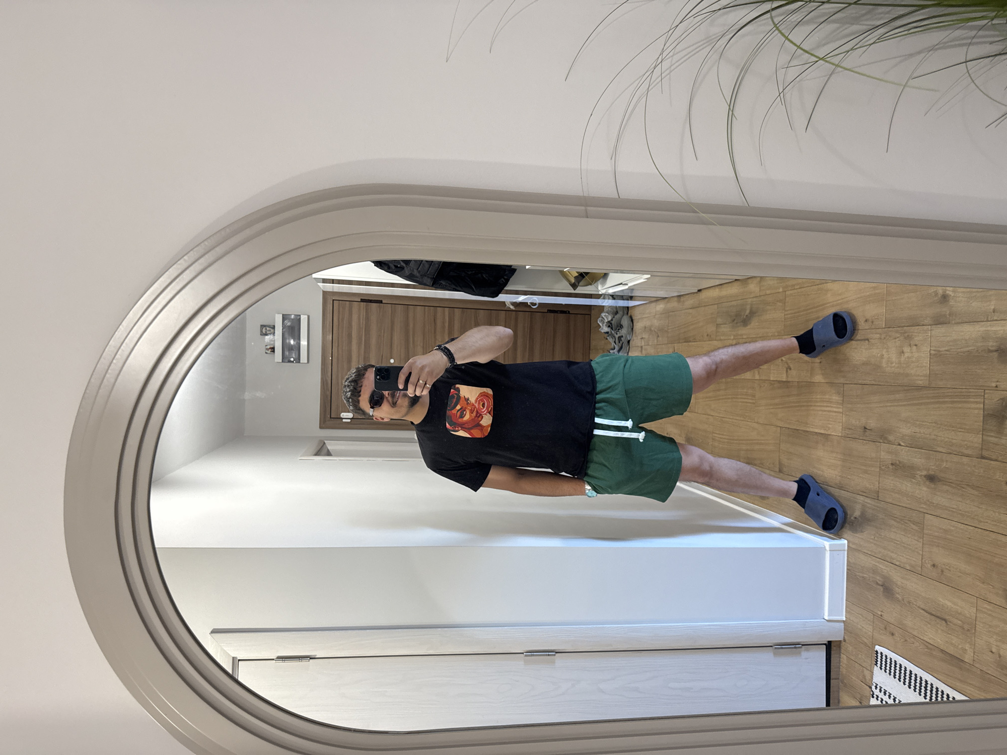 A man stands in front of an arched mirror, taking a picture of himself with a smartphone. He is wearing a black T-shirt with a graphic, green shorts, black socks, and gray slides. The room is modern with light wood flooring. There is a white door and a white vanity visible in the mirror's reflection. A plant with long, thin leaves is visible in the upper right corner of the image, outside the mirror.