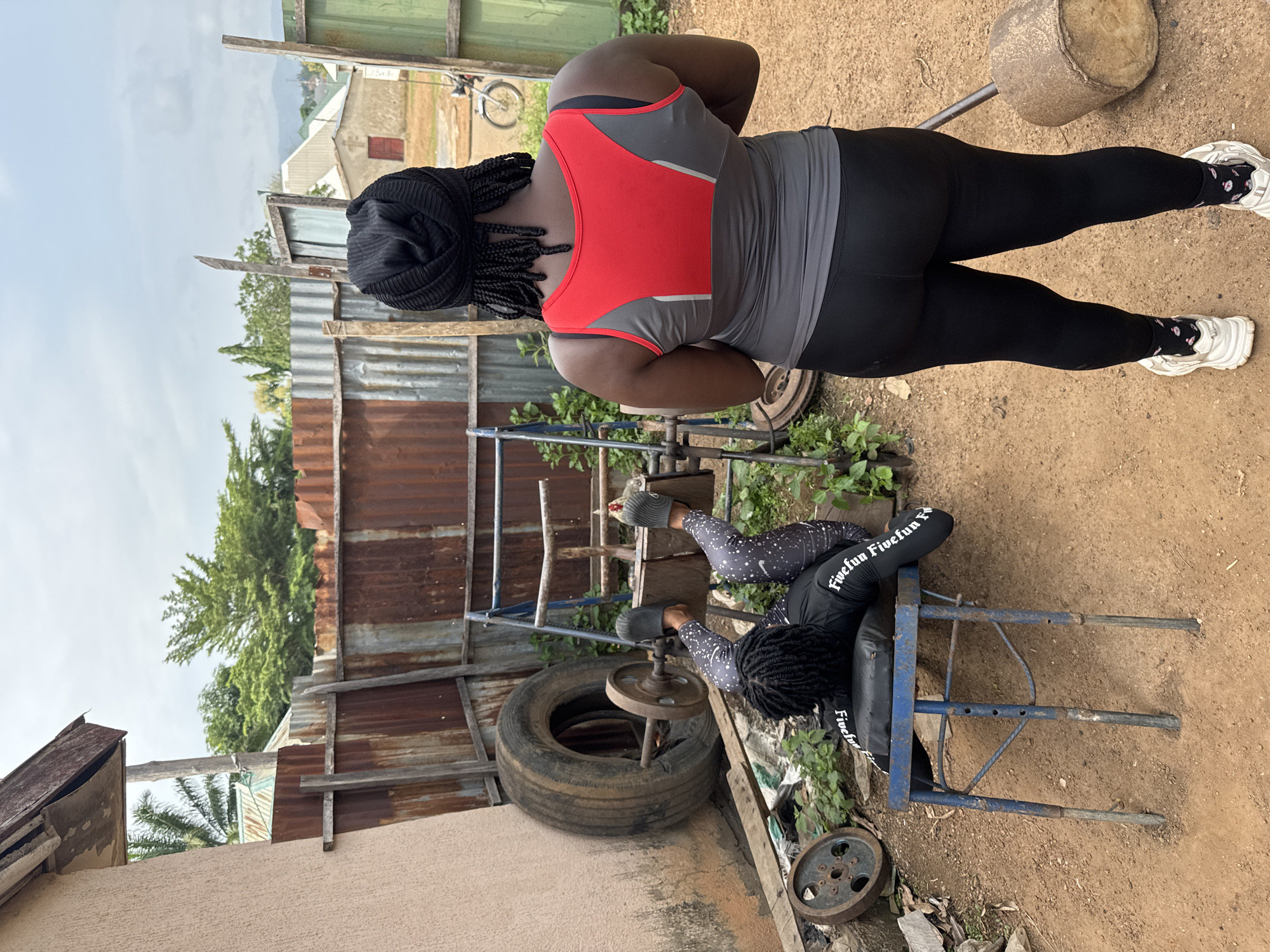 In Akwanga, Nigeria, two Black individuals are engaged in an outdoor exercise session. The first individual, wearing a black headscarf, a red and grey tank top, black leggings, and white sneakers, stands facing away from the camera, observing the second individual. The second individual, lying on a blue exercise bench with their legs positioned on a wooden apparatus, is performing a leg press exercise with a makeshift weight, which appears to be a tire with a metal rod and plates. They are dressed in a black long-sleeved shirt with white text and patterned leggings. The environment is somewhat rustic, with a corrugated iron fence in the background, along with some overgrown vegetation and scattered debris, suggesting a makeshift outdoor gym. The sky is overcast.