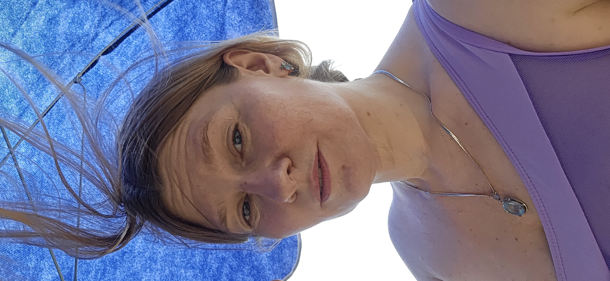 A close-up, head-and-shoulders shot of a fair-skinned woman with blonde hair is taken outdoors. She is looking directly at the camera with a neutral expression, her hair is windswept and partially obscured by a large blue umbrella overhead. She is wearing a light purple swimsuit and a silver necklace with a pendant. A small earring is visible on her right ear. The background is a white sky. The woman appears to be in a coastal area, possibly near the city of Dyurso, Russia, given the bright natural light and outdoor setting. The focus is on her face and upper body, with the environment serving as a backdrop.