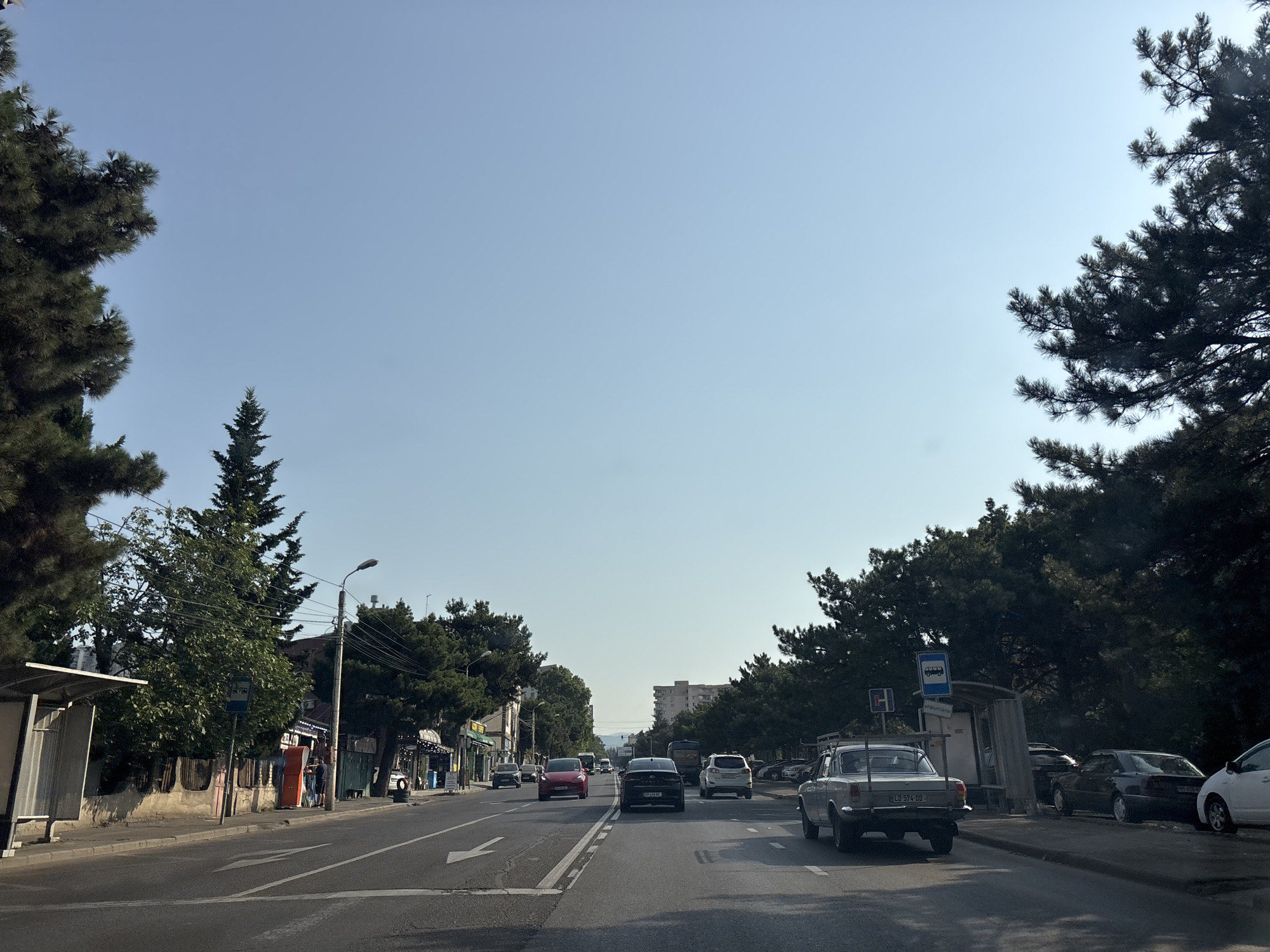 The image captures a street scene in Tbilisi, Georgia, under a clear sky. The perspective is from a vehicle moving along a multi-lane road bordered by trees and buildings. On the left side of the road, a bus stop shelter is visible, along with shops and houses. The right side of the road features parked cars and more trees. Several cars are in motion on the road, including a white older model car in the foreground on the right, and a black car in the center lane. The environment appears to be a well-established urban area with a mix of residential and commercial properties.