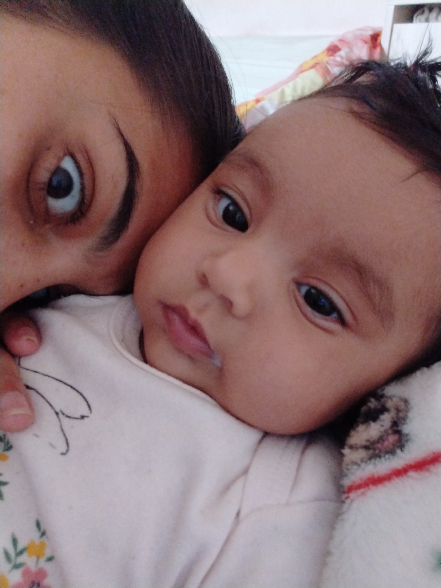 A close-up shot features a woman and a baby. The woman's face, specifically her eye and eyebrow, are visible in the upper left corner. The baby lies on a white surface, possibly bedding, wearing a light-colored onesie with floral and rabbit designs. A small amount of white liquid is visible near the baby's mouth. A blanket or padded surface is positioned beneath the baby's head. The location is unknown.