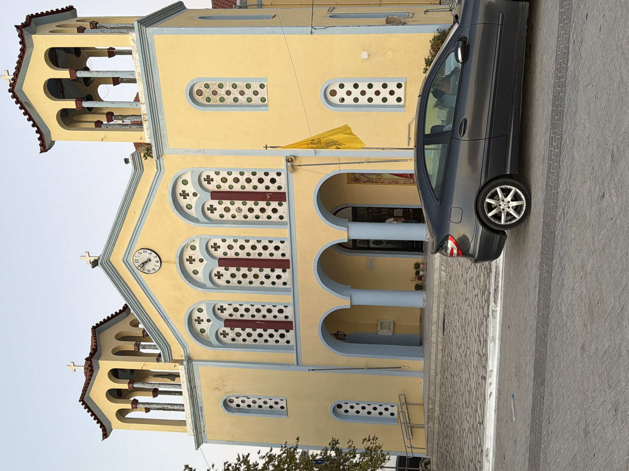 The image depicts the exterior of a yellow church in the Municipal Unit of Alyzia, Greece. The church features a prominent clock on its facade and two bell towers. A dark grey coupe is parked in front of the church. A yellow flag with an emblem is flying from a pole to the right of the entrance. The ground in front of the church is paved with stone tiles. The sky is clear and a light blue. The overall scene suggests a peaceful, urban setting.