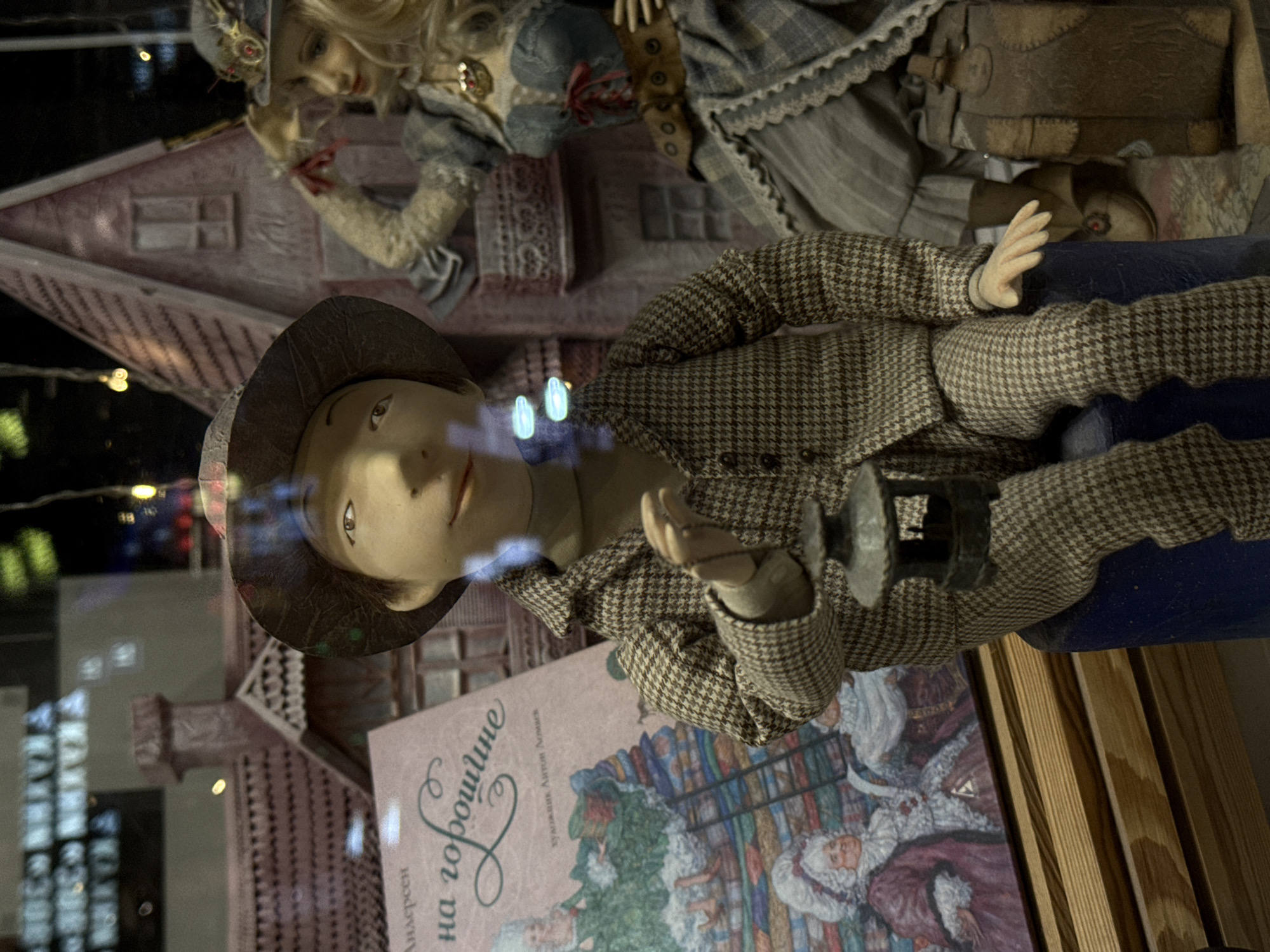 The image depicts a store window in Moscow, Russia. Inside the window, there are two dolls and a book, along with a pink painted dollhouse. The lower doll wears a houndstooth suit and a hat. The upper doll is blonde and wears a blue and grey dress with a brown bag next to it. The book's cover is visible, it features colorful illustrations. There are wooden boards in the right of the scene. Bright reflections can be seen across the window's surface.