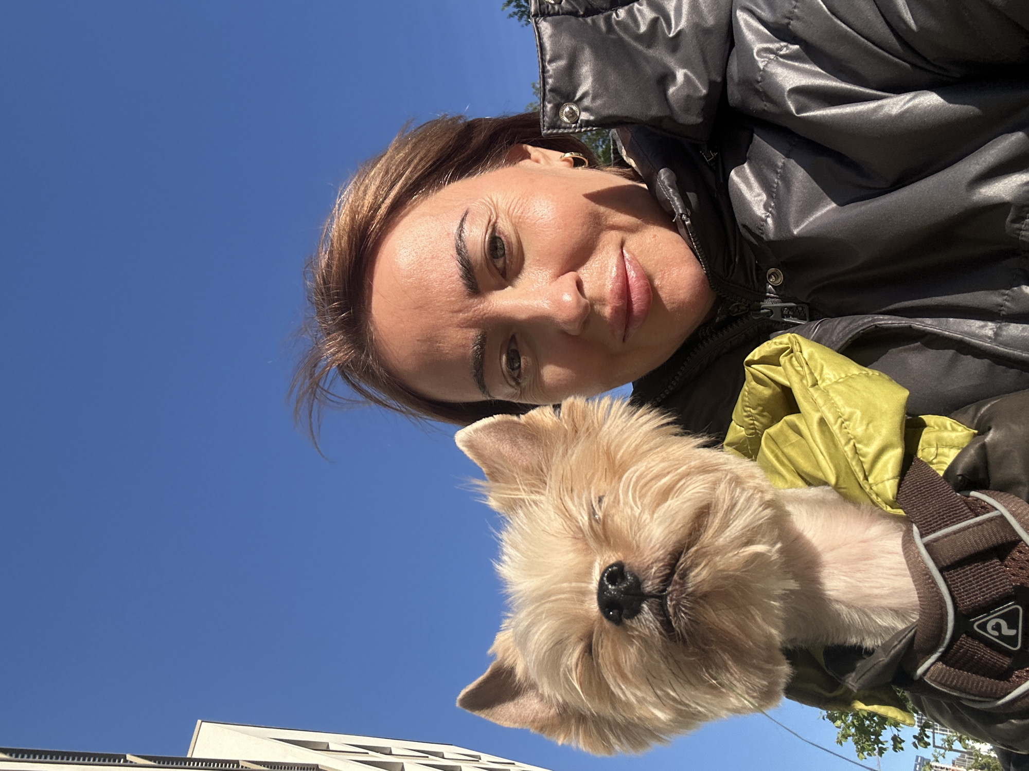A woman and a small dog are positioned in the foreground of an outdoor photograph taken in daylight under a clear blue sky. The woman, positioned towards the top, is wearing a shiny, dark puffer jacket with a partially visible zipper. She has light-toned skin and light brown hair and is looking at the camera with a slight smile. Below her, the dog, with light brown fur and a black nose, appears to be in a harness, also looking towards the camera.
In the background, the bottom of a building with white, horizontal features is visible, suggesting an urban setting. Specific details linking the image to Yekaterinburg, Russia are not evident in the photograph. No distinct landmarks or signage can be identified to definitively place the location.