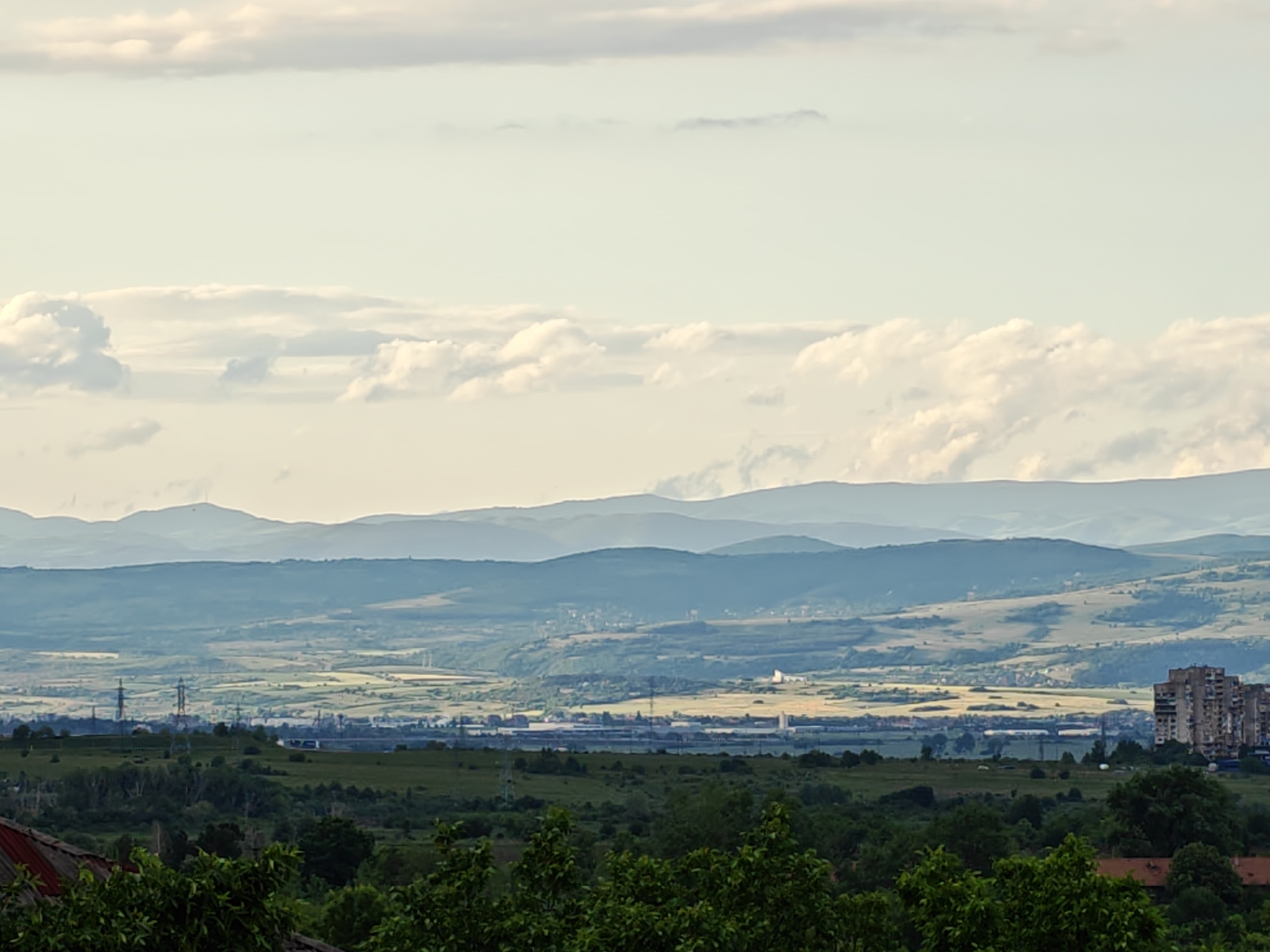 This is a panoramic landscape photograph showcasing a wide valley or plain flanked by multiple layers of hills and mountains receding into the hazy distance, with a clear sky overhead. The scene is likely viewed from an elevated position.
The upper third of the image is dominated by a light sky filled with scattered, fluffy cumulus clouds, suggesting fair but slightly overcast weather. Below the sky, several distinct ranges of mountains and hills stretch across the horizon. These layers exhibit atmospheric perspective, appearing progressively bluer, greyer, and more muted as they extend further away.
The expansive valley in the middle ground features a patchwork of agricultural fields, varying in shades of green and yellowish-green. Interspersed within this landscape are signs of human development, including industrial structures or large buildings, power line towers (pylons), and what appears to be a town or industrial complex stretching horizontally across the plain.
To the far right, a prominent, multi-story apartment building, constructed from concrete, stands out against the natural backdrop, suggesting an urban or suburban edge. The immediate foreground is filled with the dark green foliage of trees and shrubs, with a glimpse of a reddish-brown rooftop visible on the bottom left.
The lighting is soft and diffused, indicating a daytime scene, possibly late morning or afternoon, under a slightly hazy sky. There is no discernible activity or visible text. The location context indicates this is a view in or around Sofia, Bulgaria, consistent with the blend of natural landscapes and Soviet-era architectural elements.