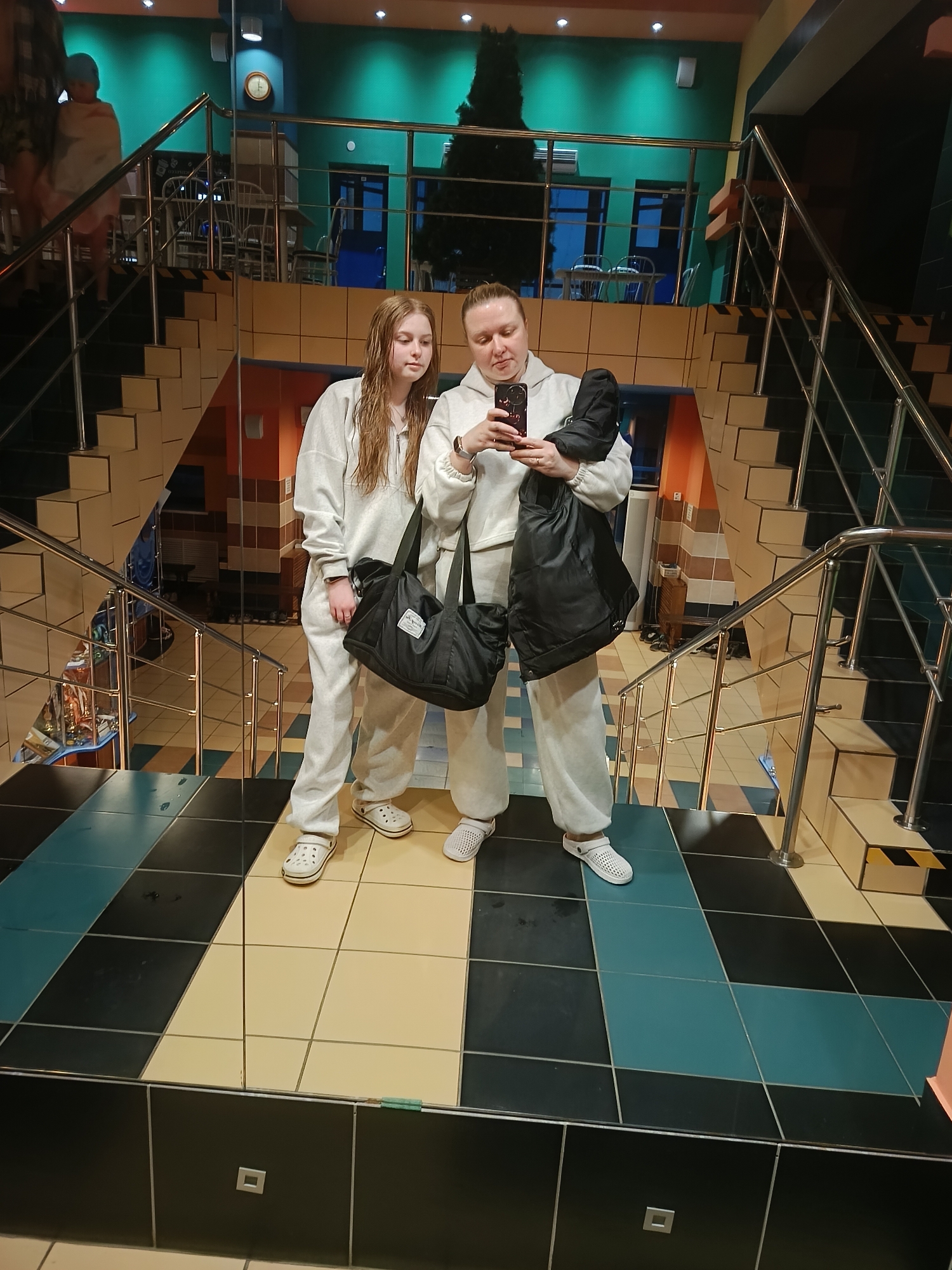 Two women stand on a tile landing, likely in a shopping mall in Chaikovsky, Russia. They both wear light gray sweatsuits and casual shoes. The woman on the right holds a black phone and a black jacket. She looks at the phone. The woman on the left looks at the phone as well. They stand in front of stairs with steel railings. The flooring and walls feature yellow, black, and teal tile. A Christmas tree stands on an upper level of the building. People walk on the stairs in the background.