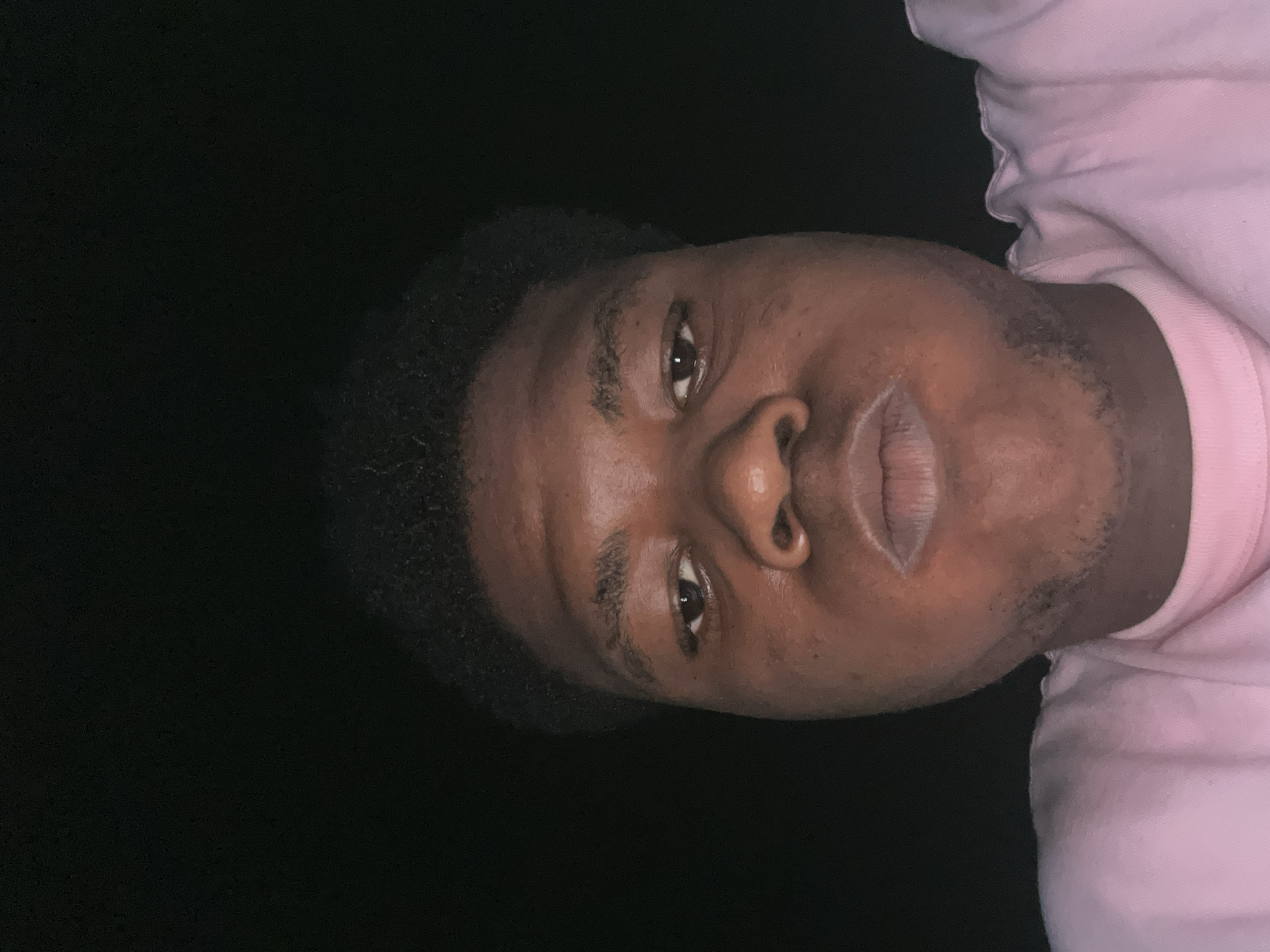 The image is a close-up, head-on selfie of a young Black male with dark skin and short, dark, curly hair. He is wearing a pink crew-neck t-shirt. His lips are slightly pursed. The background is completely black, suggesting a dark environment, possibly at night. The lighting appears to be from above and in front of the subject. The image was taken in Ihiagwa, Nigeria.