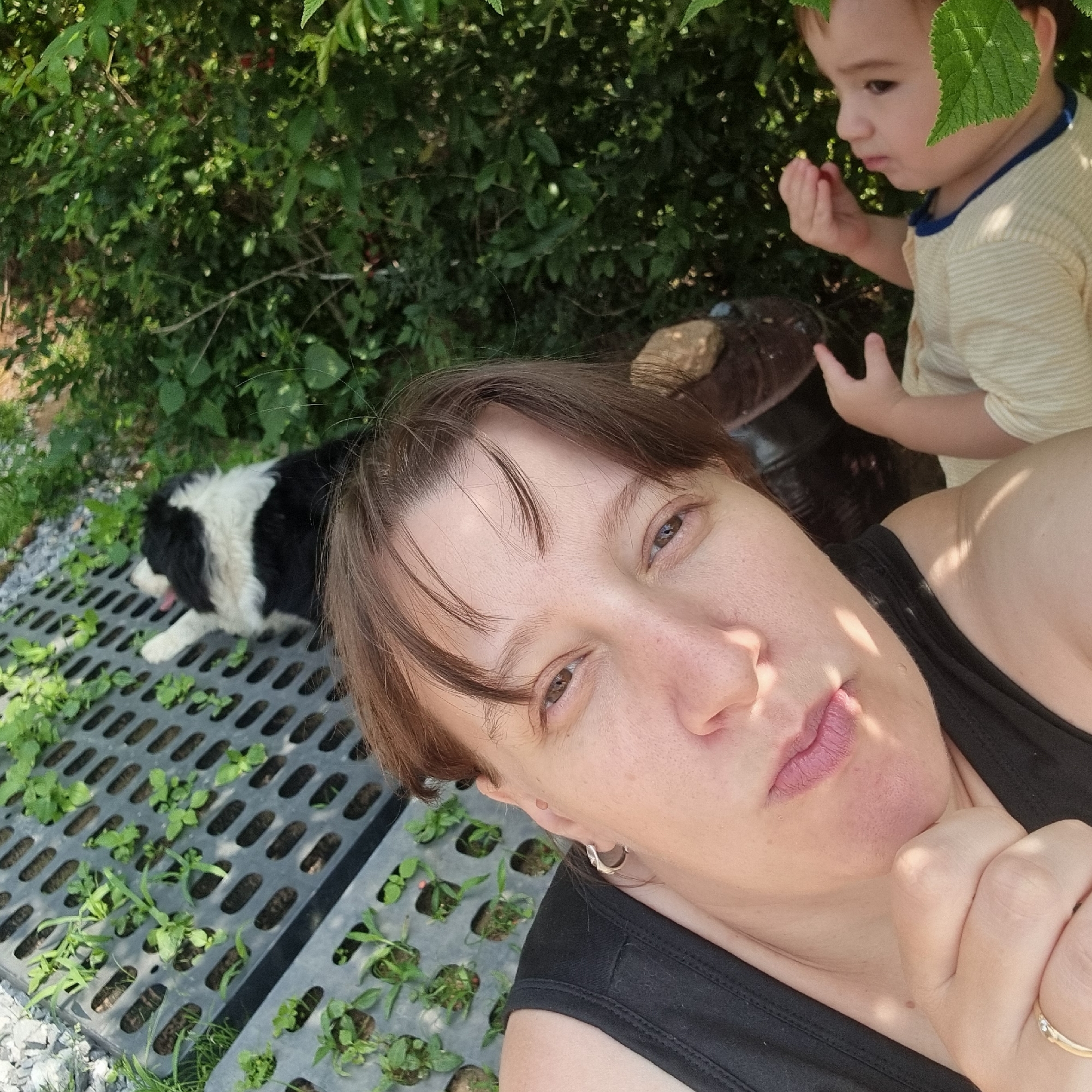 A high-angle, close-up selfie shows a woman in the foreground, looking upwards and to the left. She has brown hair, brown eyes, and is wearing a black tank top. Sunlight casts shadows on her face. To her right, a toddler in a striped shirt is reaching for something with his right hand. Behind them, a black and white dog lies on a grey plastic tray filled with small green plants. The background is filled with dense green foliage. The scene appears to be outdoors, possibly a garden or yard, with some gravel and dirt visible on the ground. The location is identified as Dunnae-myeon, South Korea.