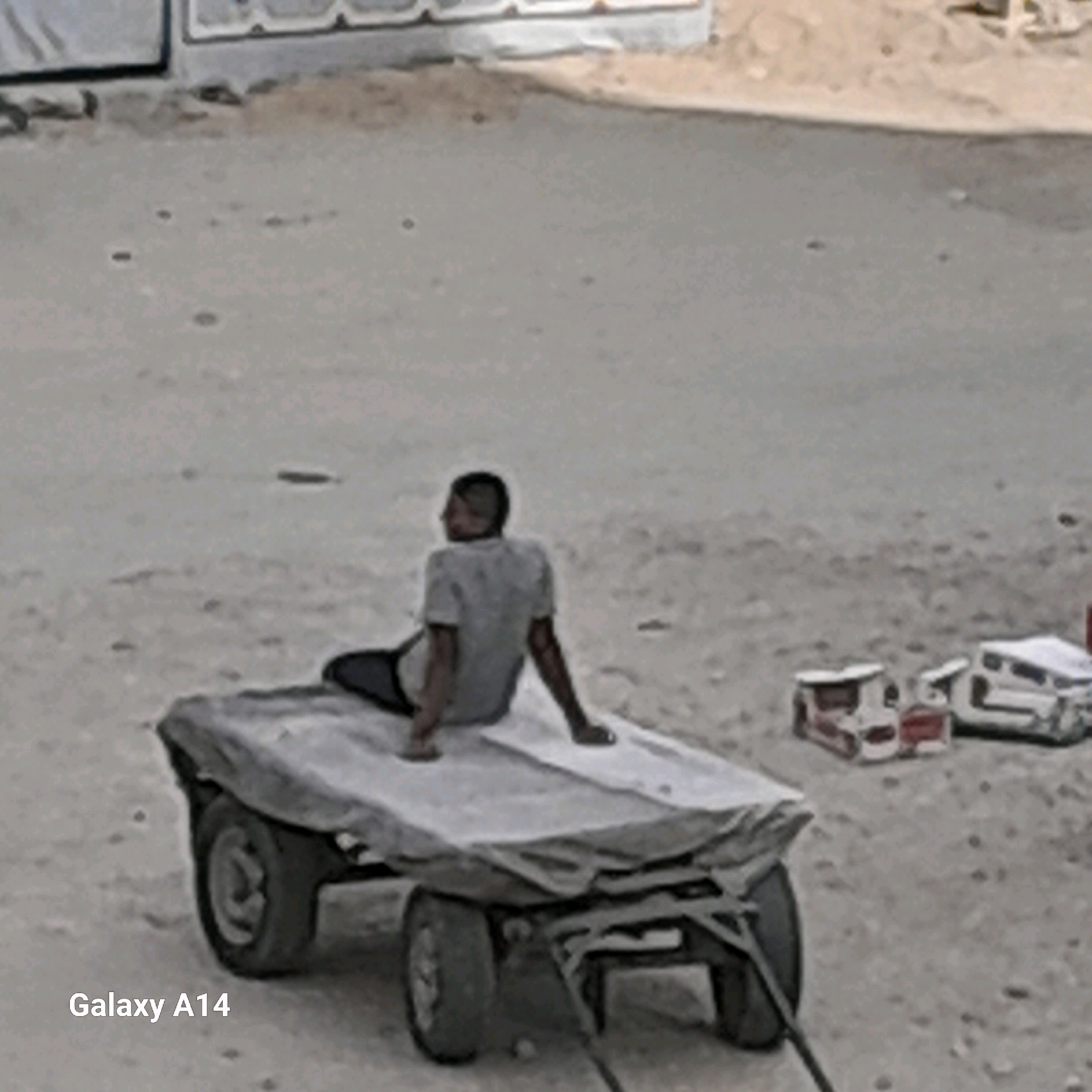 The image depicts a boy sitting on a cart in Khan Yunis, Palestinian Territories. The cart is covered with a tarp and has two wheels. The boy is wearing a grey t-shirt and dark pants, and appears to be looking away from the camera. The ground is sandy and there are some scattered objects in the background that resemble toy cars. The scene suggests a simple, possibly rural or informal setting.