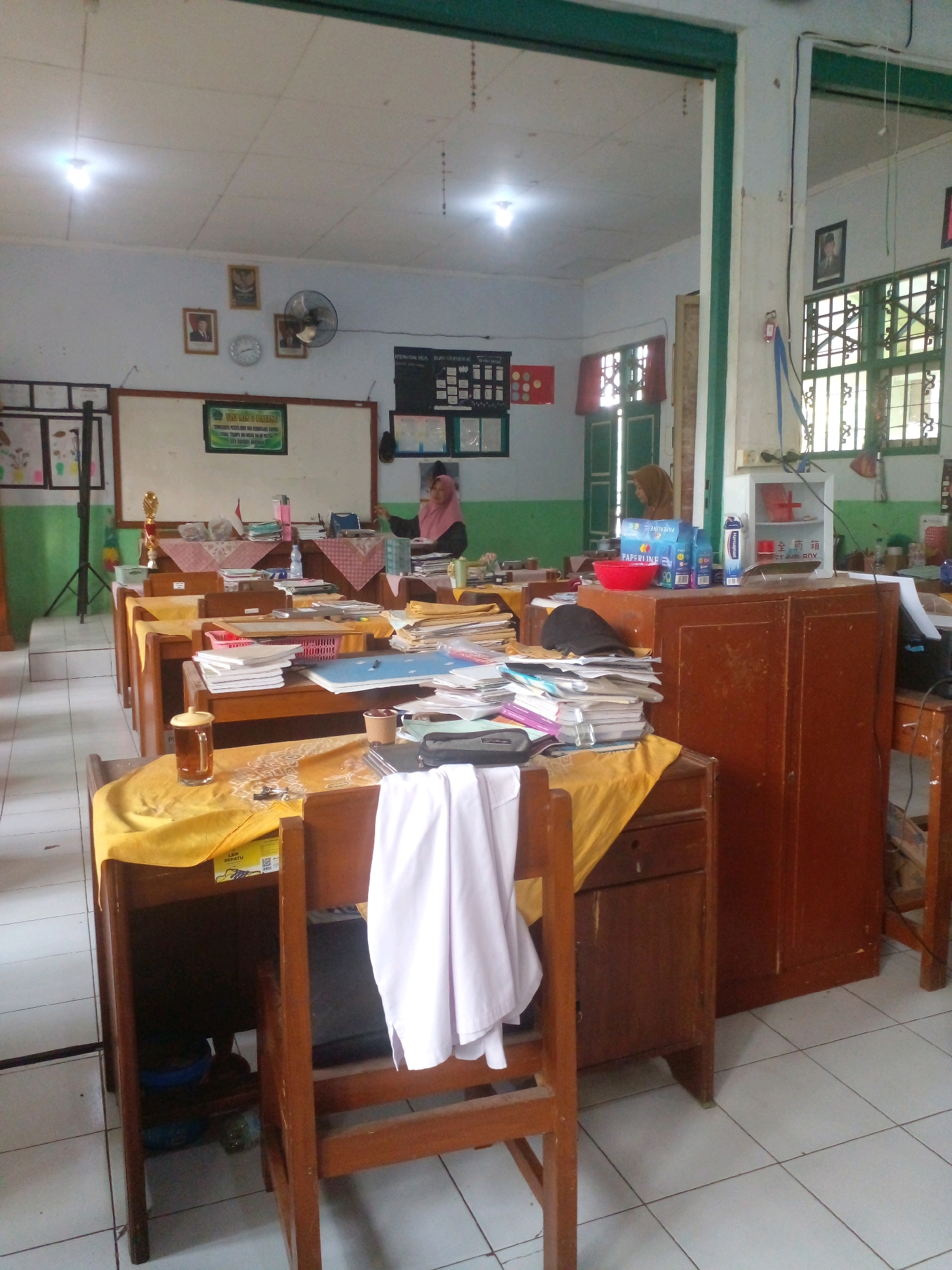 The image depicts an indoor scene, likely a classroom, with rows of wooden desks and chairs arranged in the foreground and midground. The desks are covered with yellow cloths and cluttered with various items including stacks of paper, books, a glass of tea, and a red basket. A white cloth is draped over the back of a chair in the foreground.
In the background, a whiteboard is visible on the wall, with posters and framed pictures above it. A ceiling fan is mounted on the wall to the right of the whiteboard. A woman wearing a hijab is standing near the whiteboard. Further to the right, a large wooden cabinet with two doors stands against the wall. On top of the cabinet, a white first-aid box and several boxes of stationery are placed. The room has tiled floors and fluorescent lighting fixtures on the ceiling. Windows with green frames are visible on the right side of the image, letting in natural light.
The image was taken in the city of Banyuputih, Indonesia. The overall impression is that of an active or recently used classroom, possibly at a school.