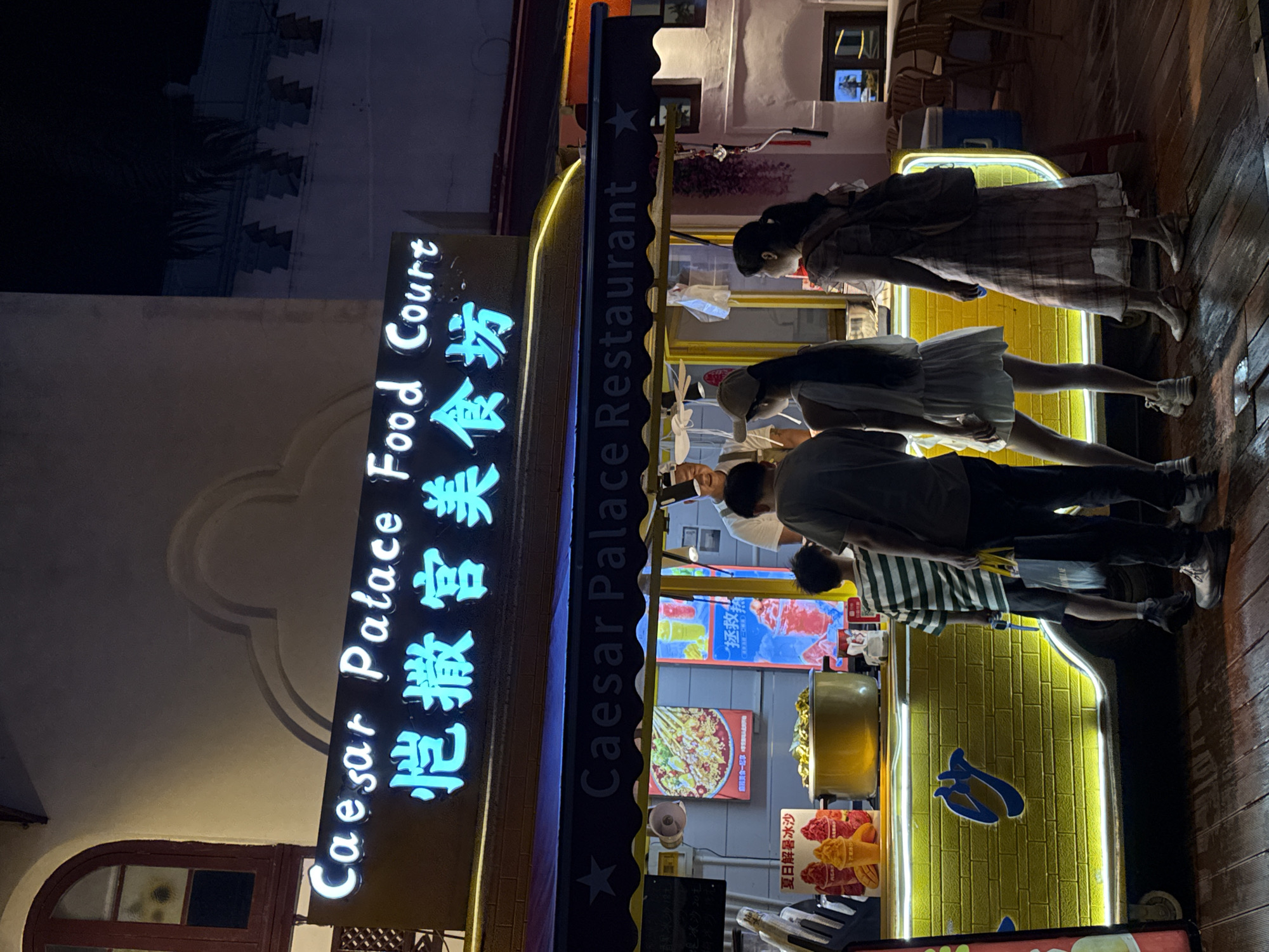 This is a night time photograph of an outdoor food stall in Nanshan District, China. The stall has a bright yellow facade with a blue logo and a blue neon sign that reads "Caesar Palace Food Court" in English and Chinese. Above the food stall, there is a dark awning with "Caesar Palace Restaurant" written on it. There are four people in front of the stall. A man, presumed to be the vendor, is standing behind the counter and interacting with the customers. Three customers are visible: two women and a young boy. One woman is wearing a white dress and a baseball cap, and the other is wearing a plaid dress and a backpack. The young boy is wearing a striped shirt and shorts. The ground is made of dark wood planks, and there is a hint of ambient light from the surrounding buildings and the food stall itself. The overall atmosphere is that of a busy night market or street food vendor.