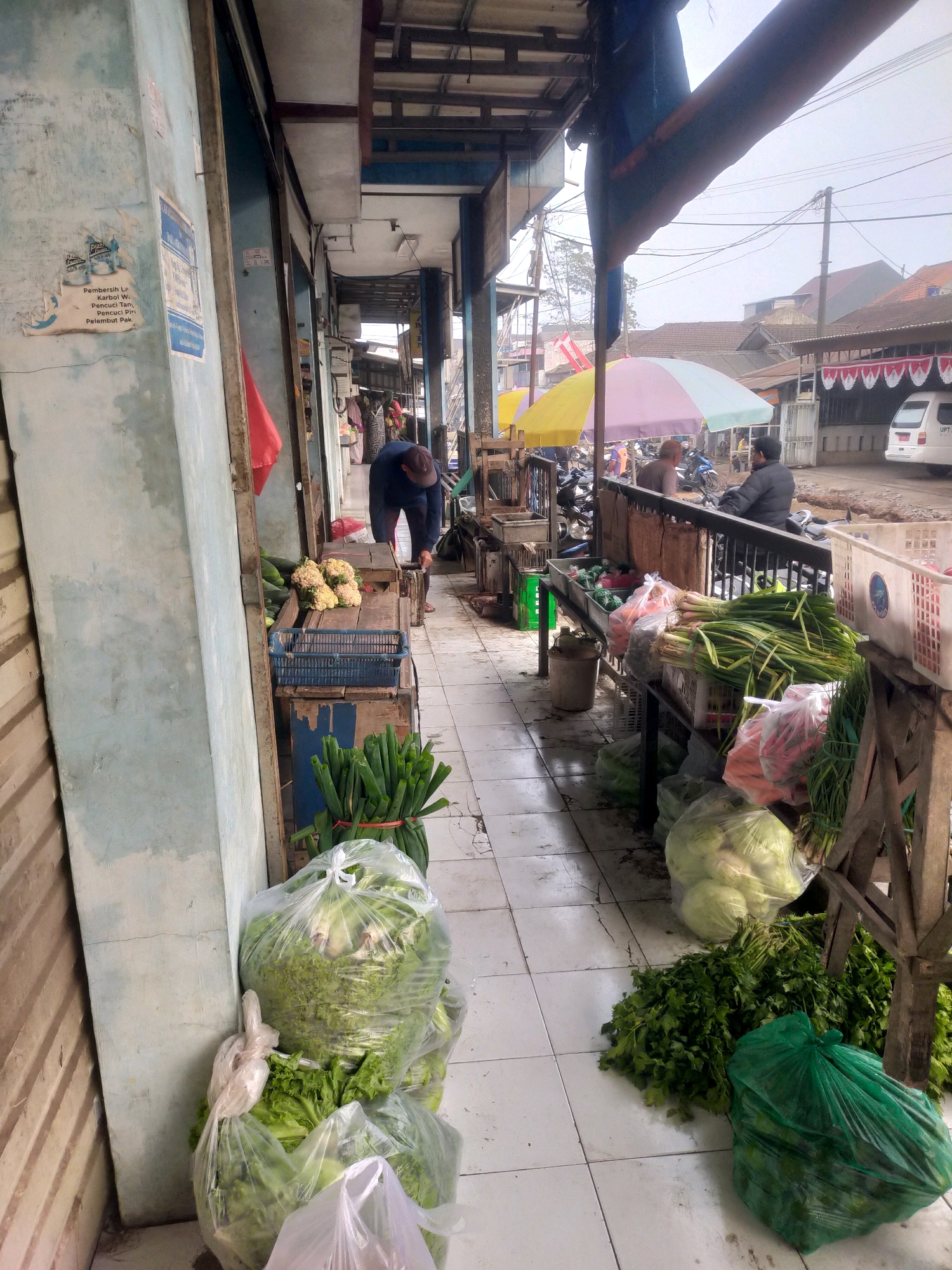 The image depicts an outdoor market stall in Bululawang, Indonesia. The scene is filled with fresh produce, primarily vegetables, displayed on wooden tables and in plastic bags. In the foreground, large plastic bags contain leafy green vegetables like lettuce. Bunches of green onions are tied with red elastic bands. Further back, cauliflowers are visible in a crate, alongside cucumbers. Various other vegetables, including carrots, are arranged on tables. A man in a blue shirt and a brown cap is bending over, appearing to be working at one of the stalls. Other people are visible in the background, some sitting or standing near motorcycles. Colorful umbrellas are set up, providing shade. The market appears to be situated along a street with buildings and electrical wires visible in the distance. The ground is tiled.