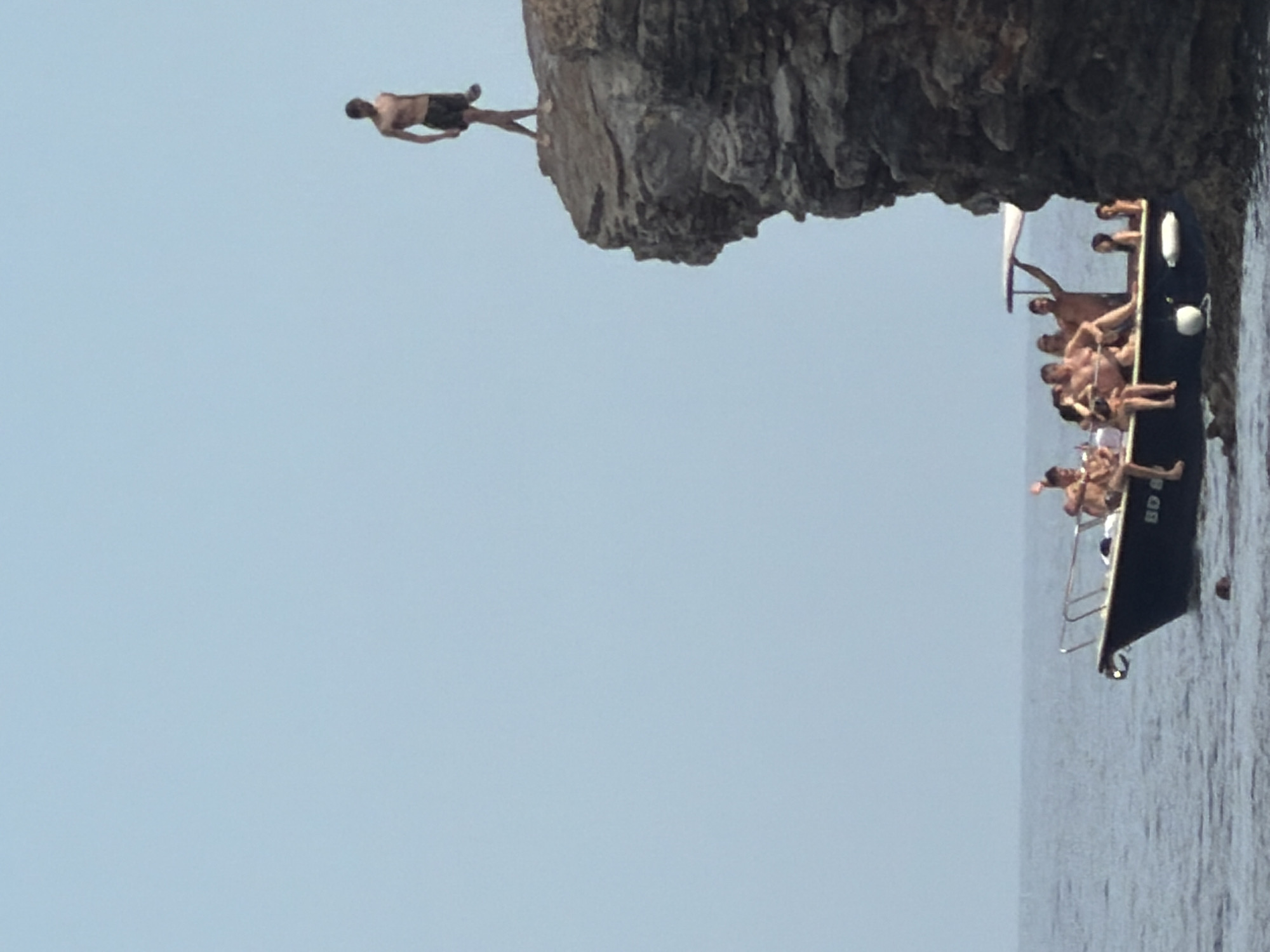 A group of people are on a boat in the water, near a rocky cliff face. On top of the cliff, a lone person is standing. The boat is dark blue with a white stripe and some people are sitting on the edge. The water is calm and the sky is clear. The scene appears to be in Budva, Montenegro, a popular coastal town.