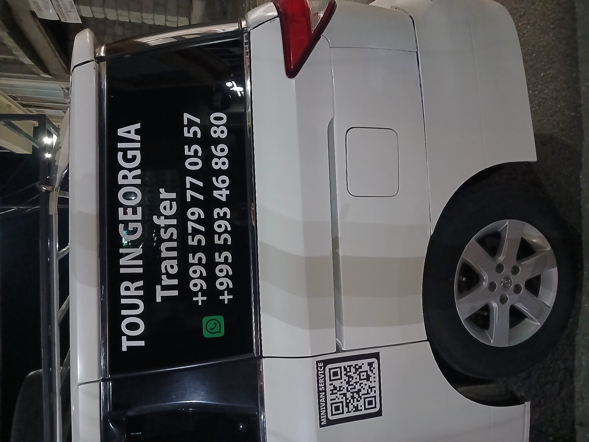 The image is a close-up, eye-level shot of the rear side of a white minivan parked outdoors at night. The minivan has a dark tinted rear window with white lettering. The text on the window reads "TOUR IN GEORGIA Transfer" and two phone numbers: "+995 579 77 05 57" and "+995 593 46 86 80". A green WhatsApp icon is visible to the left of the phone numbers. A small square QR code sticker with the text "MINIVAN SERVICE" is affixed to the lower left of the van's side panel. The background consists of a dimly lit brick wall and some foliage. The overall scene suggests a transportation service operating in Georgia. The provided metadata confirms the location as Telavi, Georgia.