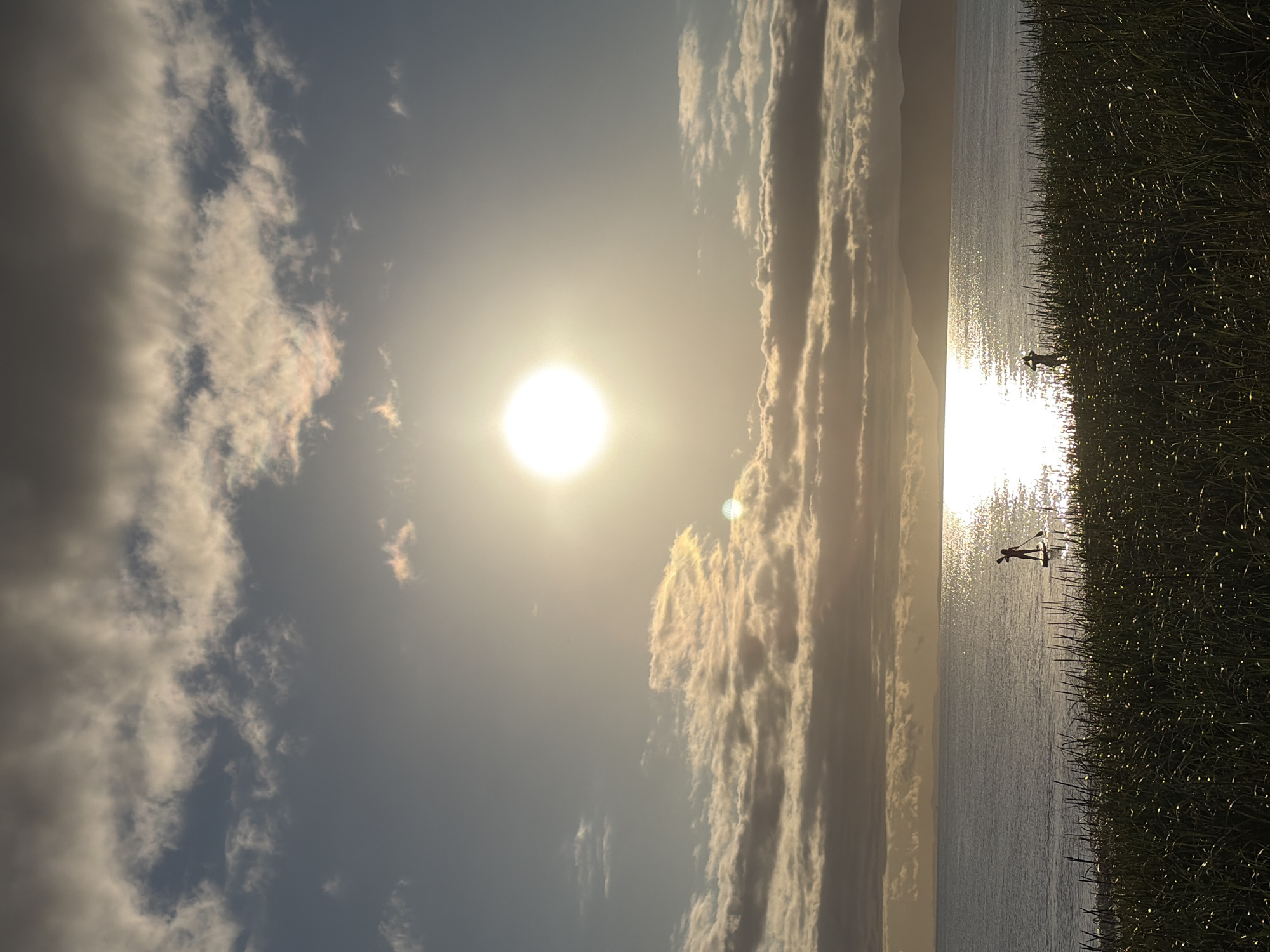 The image captures a serene coastal scene at sunset. The sun, a bright orb, is positioned high in the sky, casting a brilliant, shimmering reflection on the calm water. Silhouetted figures are visible against the glare of the sun on the water: one person is standing on a paddleboard, paddling, and another figure stands on the shore, seemingly looking out at the sea. The foreground is dominated by tall, dark grass, which is illuminated by the low-angled sunlight, creating a textured, silhouetted effect. In the background, distant hills or mountains are visible against the horizon, partially obscured by the soft glow of the setting sun and the clouds. The clouds are a mix of dark and light, with patches of sunlight breaking through, creating a dramatic sky. The overall atmosphere is one of tranquility and natural beauty.
