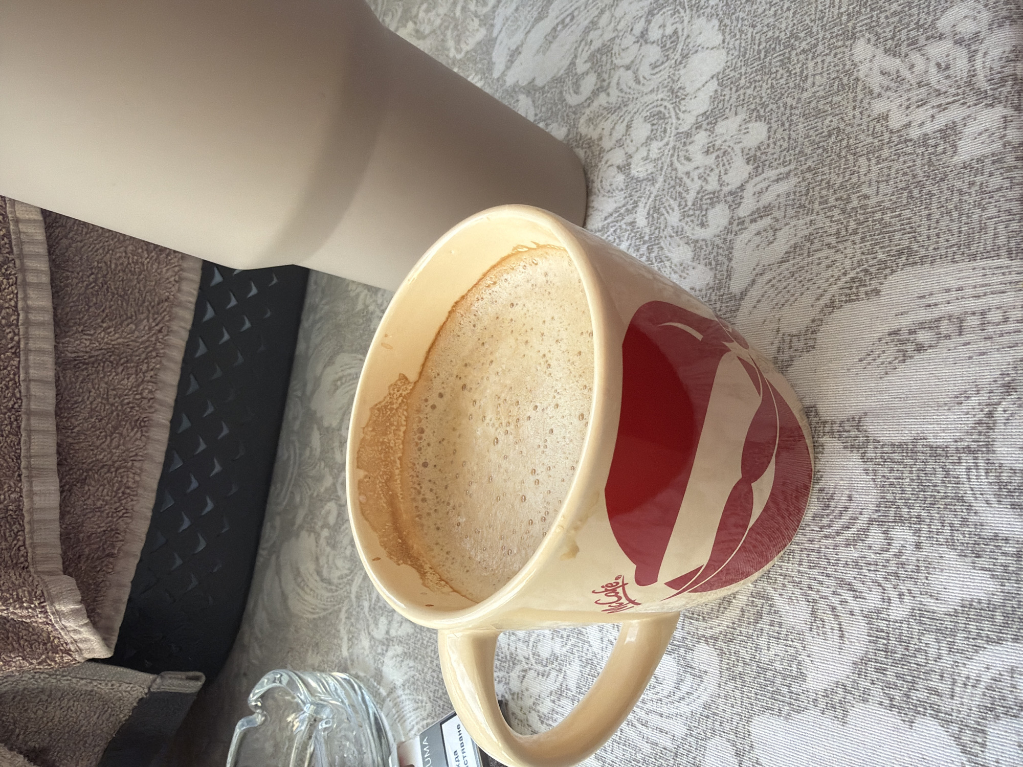 The image shows a close-up of a coffee cup with "McCafé" branding and a red lips design. The cup is filled with a frothy coffee. Next to the cup, a large, light-colored tumbler is visible. In the background, a textured brown towel is draped over a dark object. A glass ashtray and a partially visible cigarette pack are also on the table. The table is covered with a patterned tablecloth. The scene appears to be indoors, possibly on a balcony or near a window, given the lighting. There is no identifiable indication of the location being Plovdiv, Bulgaria, or any specific city.