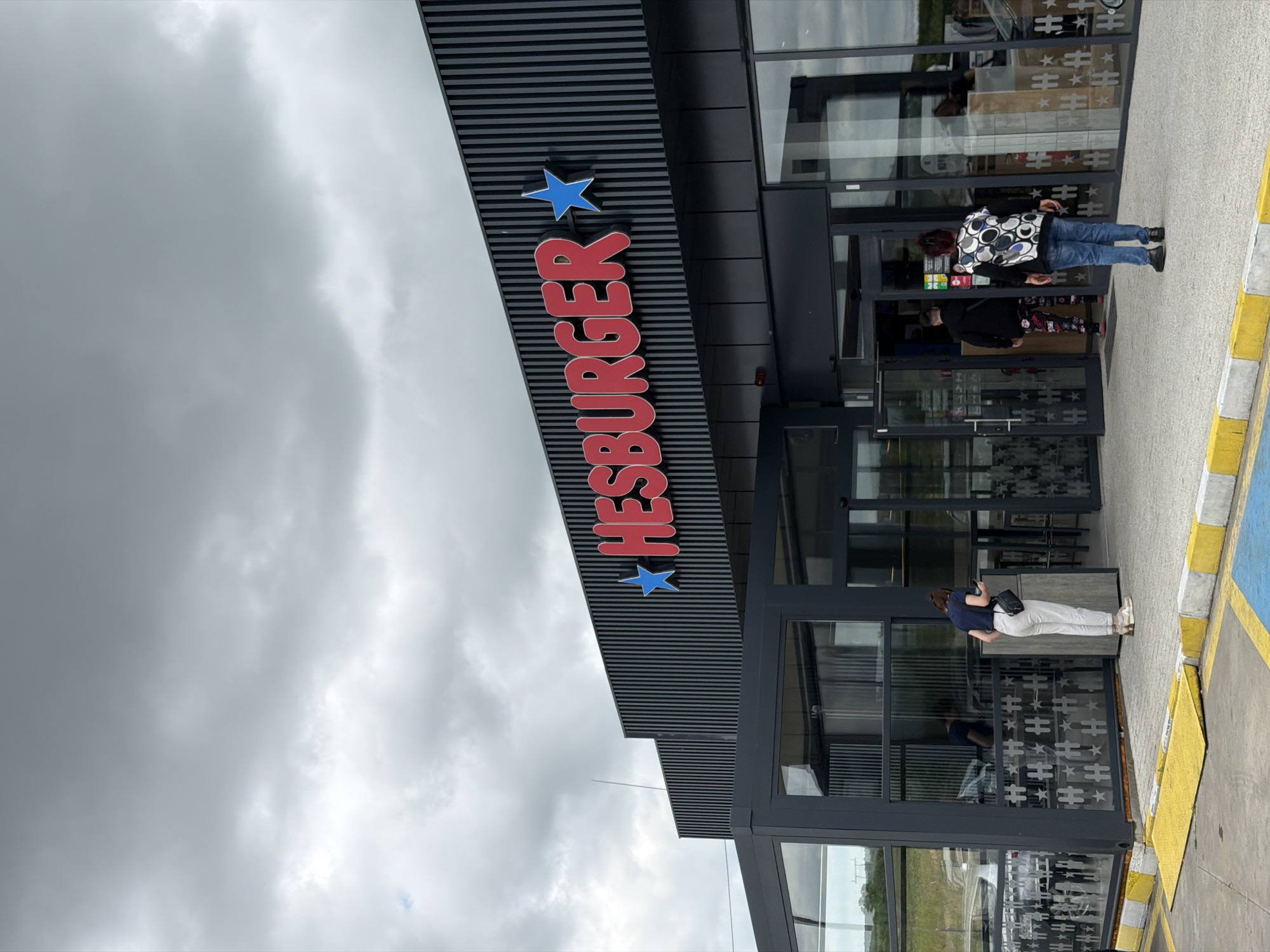 The image depicts the exterior of a Hesburger restaurant under an overcast sky. The restaurant's sign is prominently displayed with red lettering and blue stars. Several people are present near the entrance of the building, appearing to be entering or exiting. The scene is set in a parking lot, with a curb visible near the building. The reflections in the windows suggest an urban or suburban environment. I'm unable to verify the specific city.